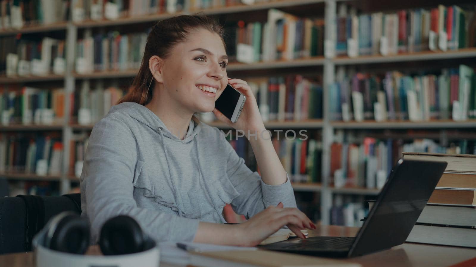 Beautiful smiling positive caucasian female student sitting at table in big library talking on smartphone looking in modern laptop and with digital tablet in front of her