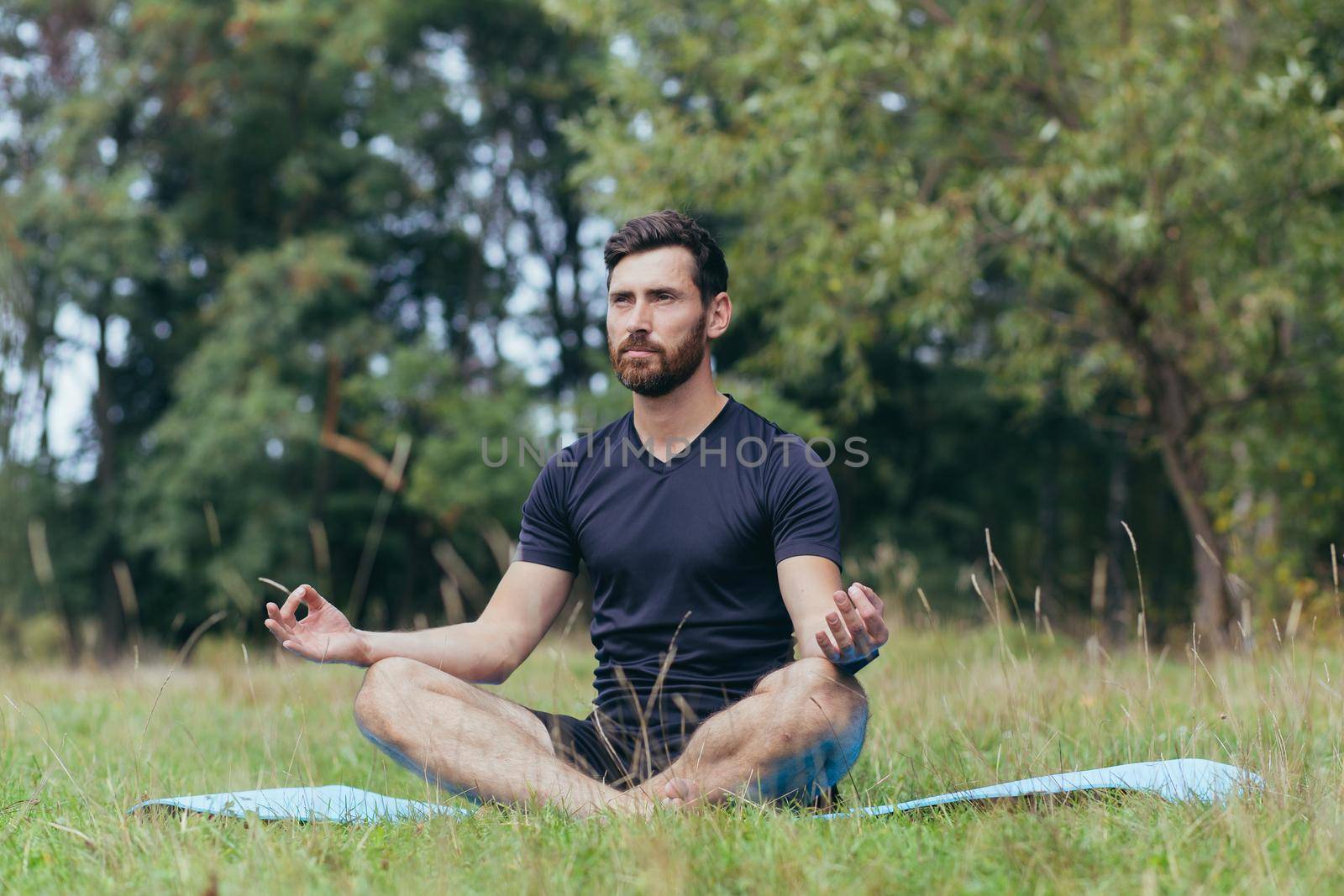 A young man with a beard sitting in the park on a mat meditates, performs exercises to improve breathing, active lifestyle