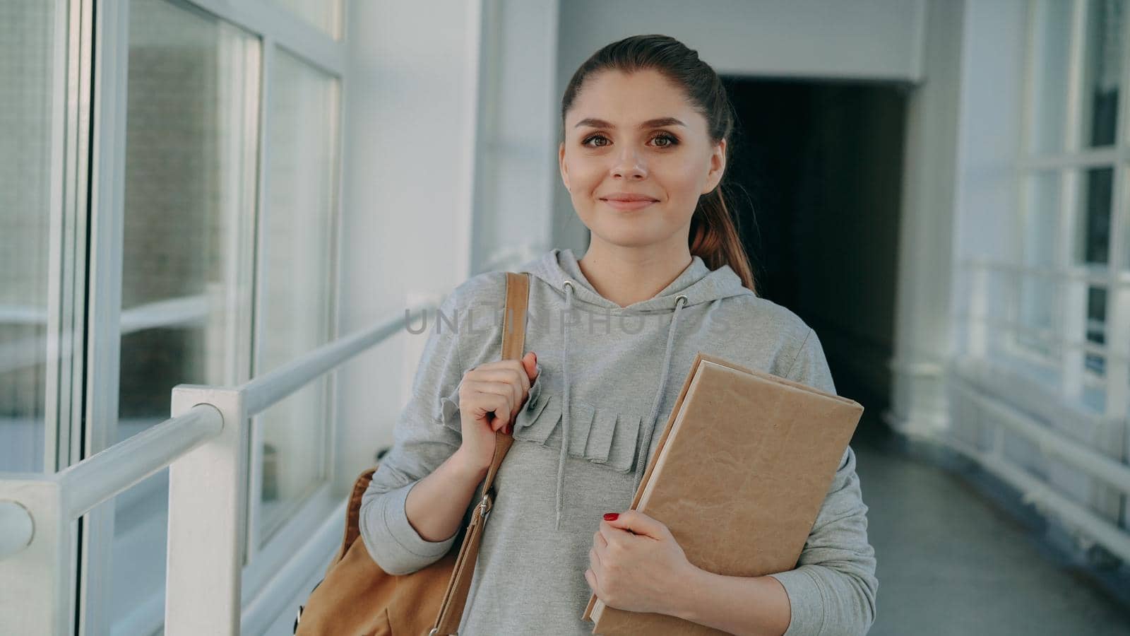 Portrait of young beautiful caucasian confident female student in casual clothing standing in wide white glassy corridor smiling positively holding textbook looking at camera