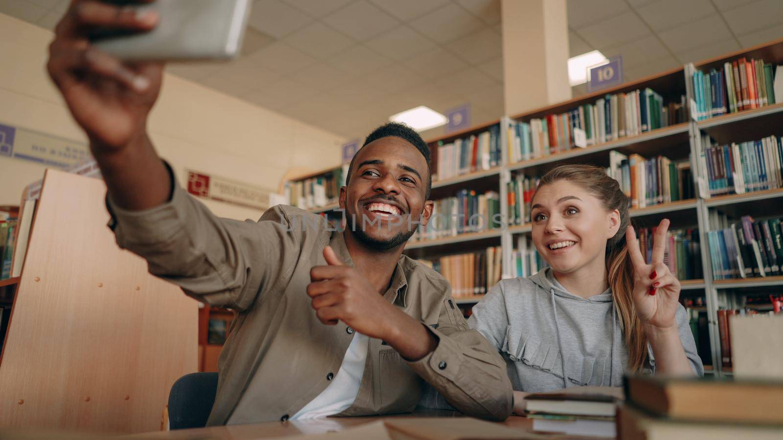 African merican guy and caucasian girl have fun smiling and taking selfie photos on smartphone camera at university library indoors. Cheerful students have rest while prepare for examination