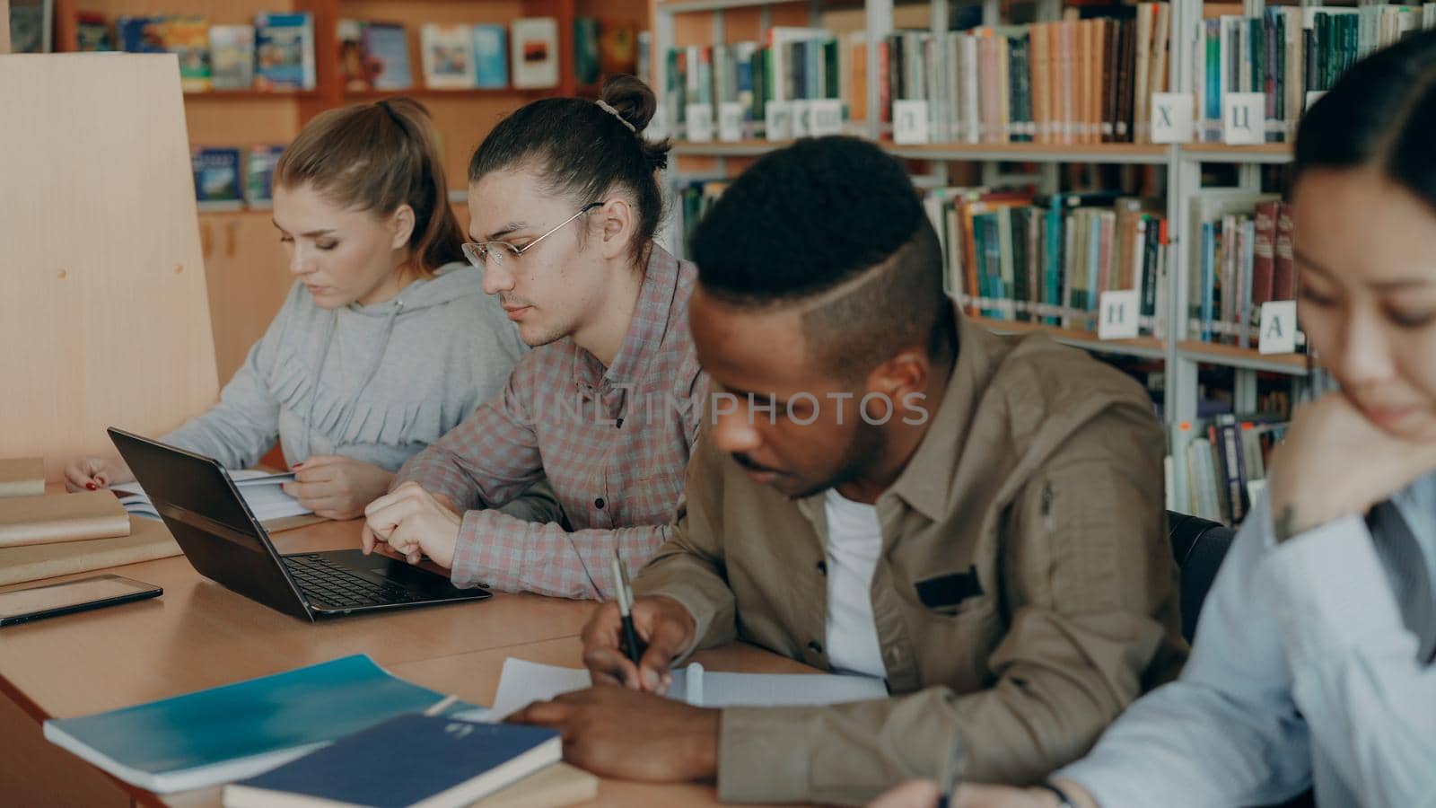 Focus on hipster male student in glasses using laptop computer while preparing for exams in university library with his classmates by silverkblack