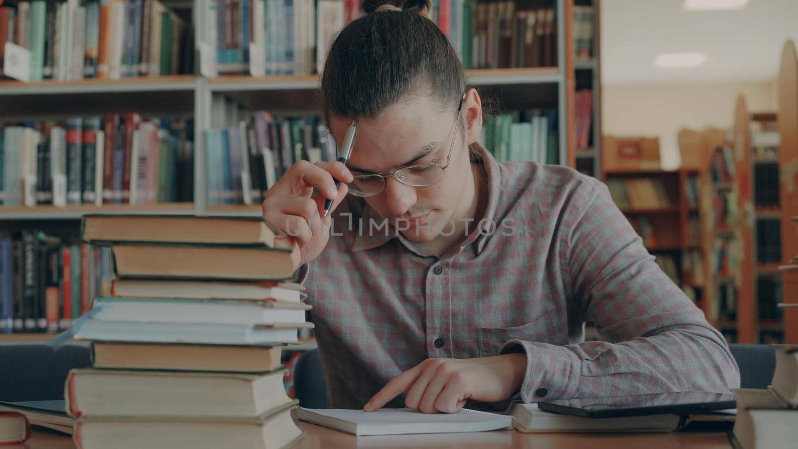 Young handsome man wearing glasses sitting at table in university library thinking over calculations in copybook. He is looking away and witing thoughtfuly by silverkblack