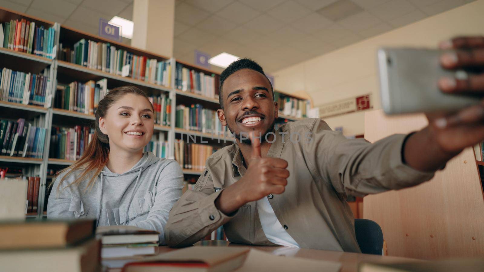 African merican guy and caucasian girl have fun smiling and taking selfie photos on smartphone camera at university library . Cheerful students have rest while prepare for examination by silverkblack