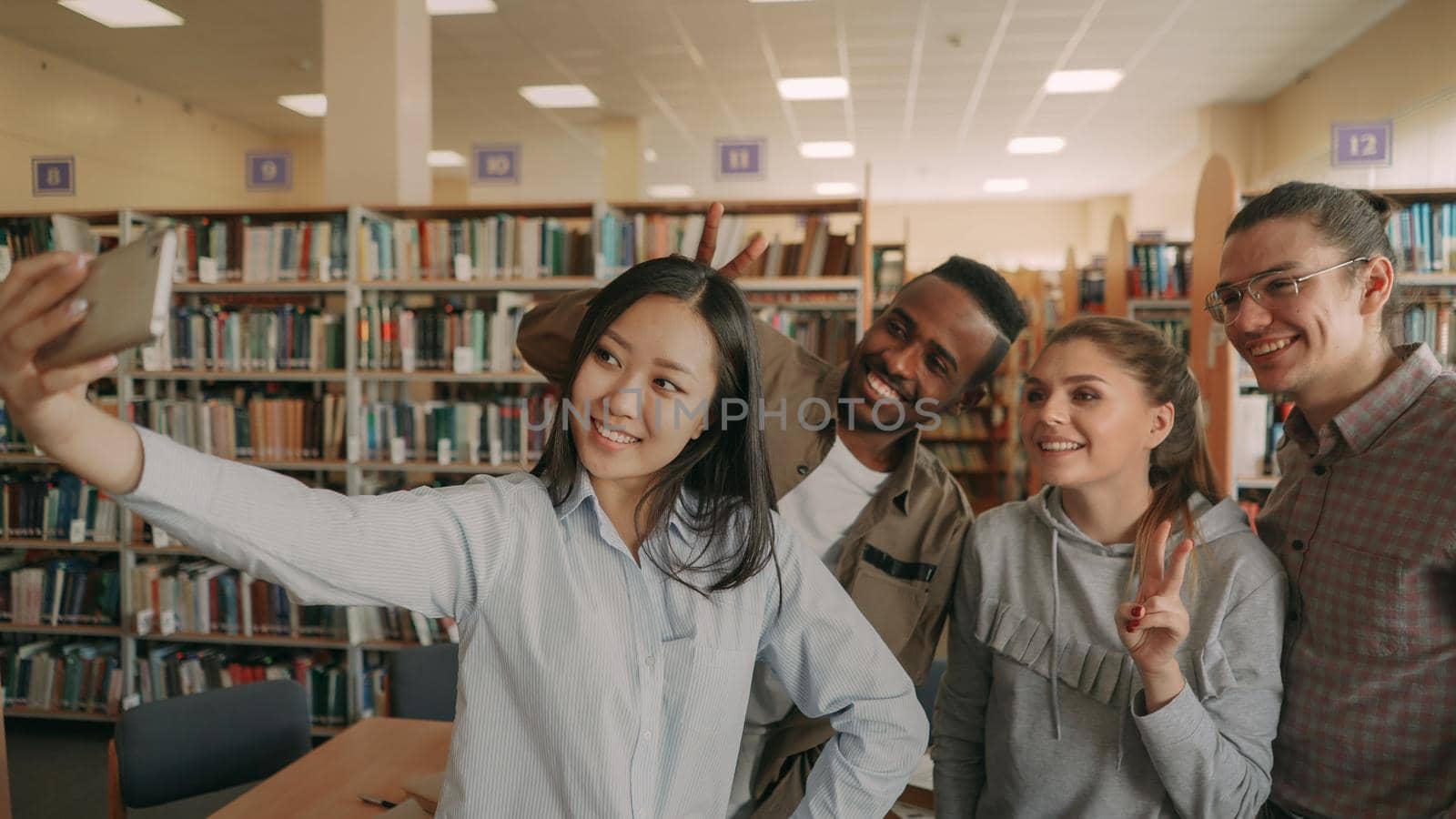 Group of international students have fun smiling and making selfie photos on smartphone camera at university library indoors. Cheerful friends have rest while preapre project together