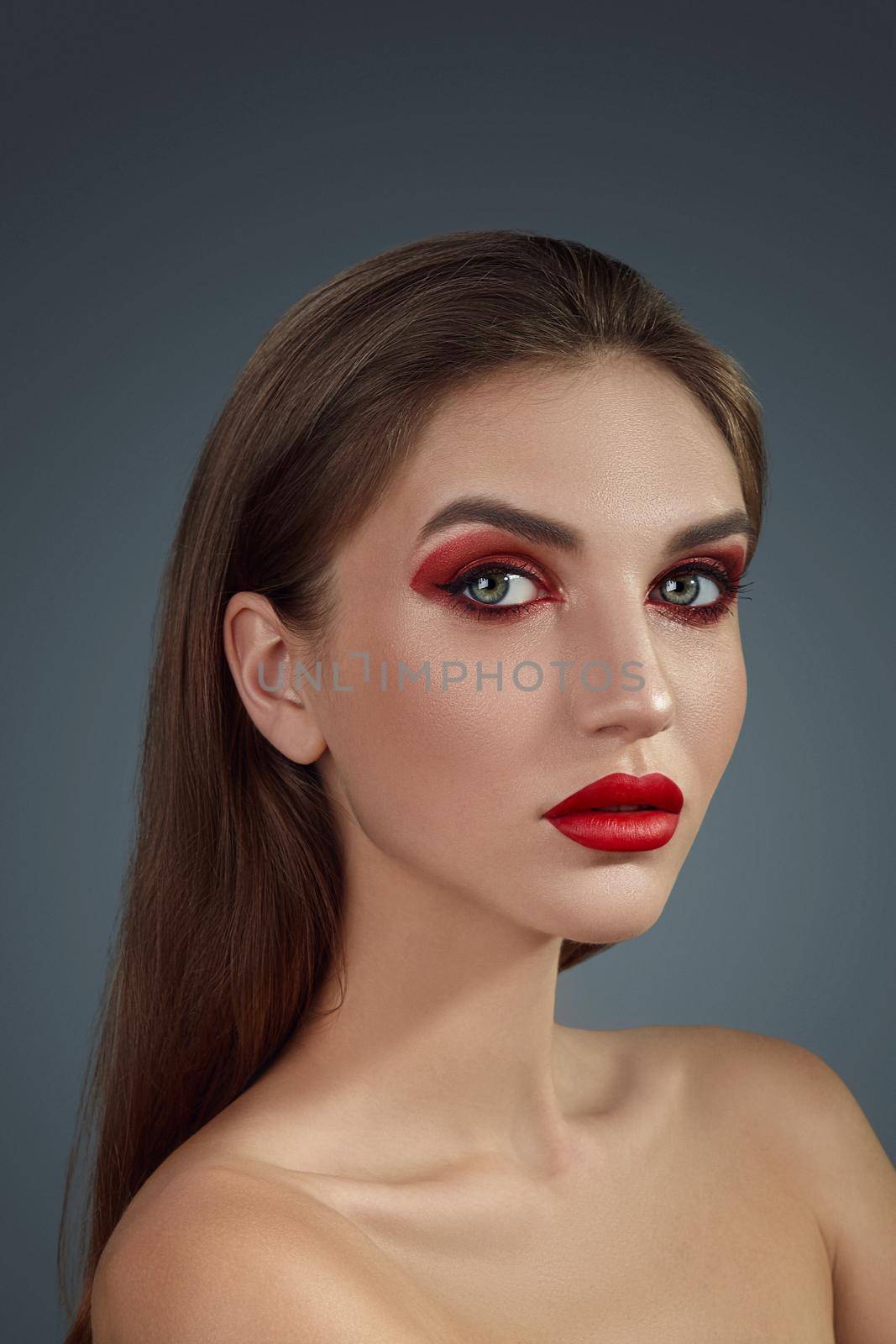 Close-up glamour portrait of a charming half-naked woman with bright smoky eyes make-up and long blond hair, looking at the camera while posing over a gray background. Fashion shiny highlighter on her skin, sexy red lips and dark eyebrows.