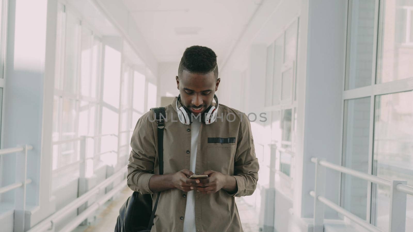 Young handsome african american male student with big white headphones walking in long lighty glassy corridor of college holding smartphone looking into it texting someone and smiling.