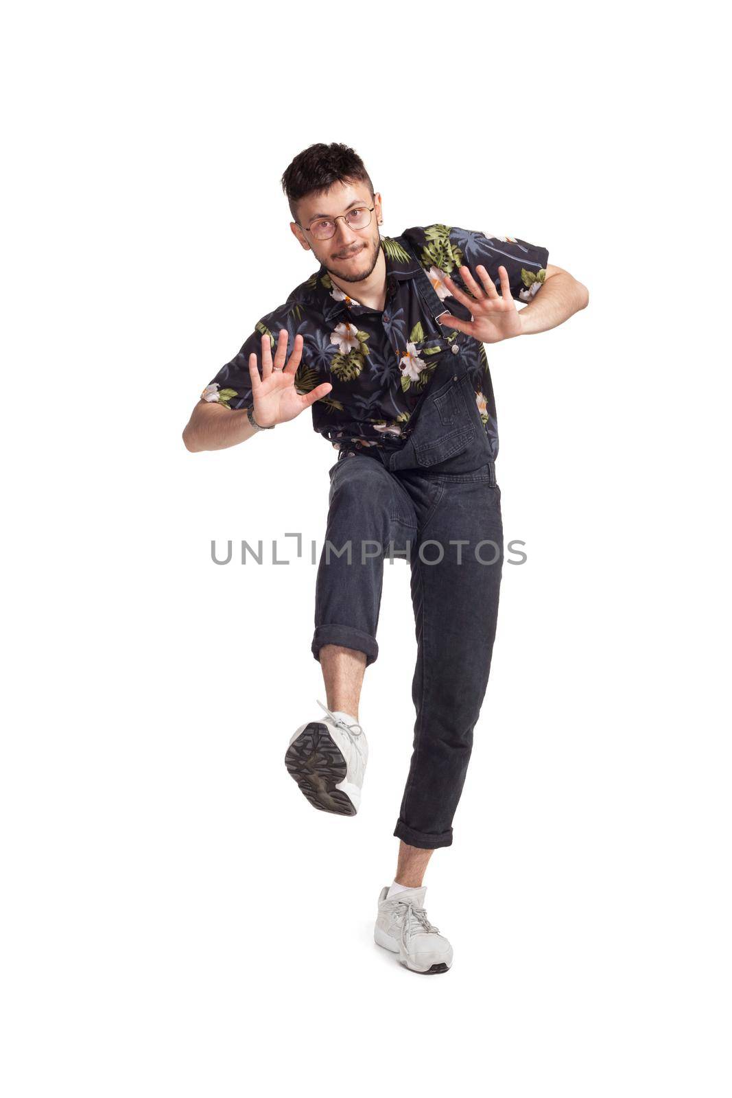 Full-length photo of a handsome dancer in glasses, black jumpsuit, funny dark t-shirt and sneakers fooling around in studio. Indoor photo of a man dancing isolated on white background. Music and imagination.