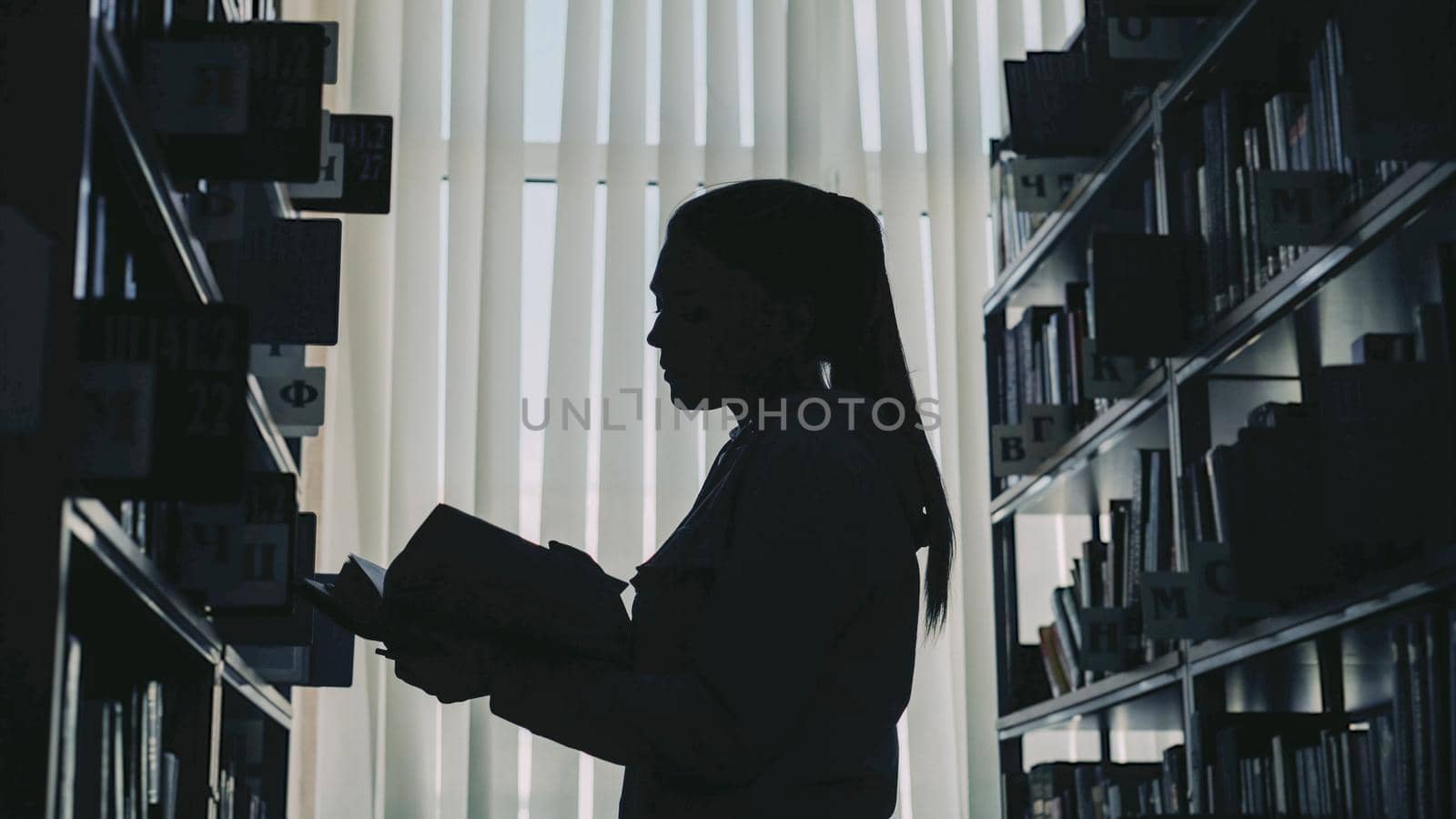 Silhouette of young student girl standing among bookshelves in big library against window holding book turning over pages and reading by silverkblack