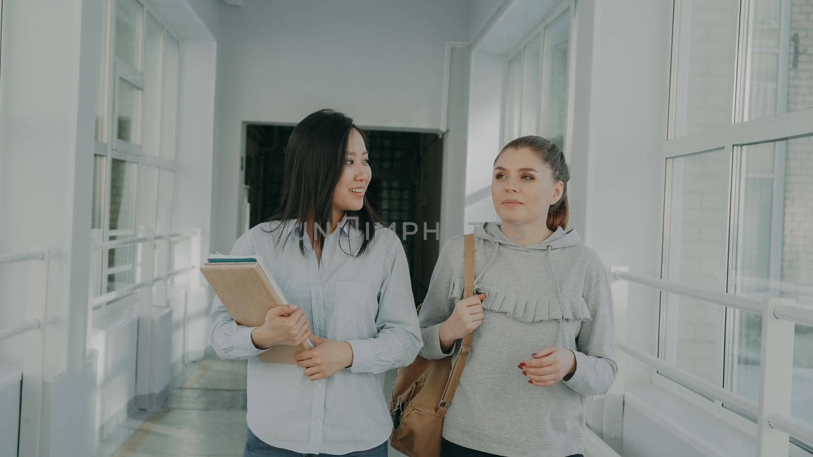 Two young beautiful female students walking in wide lighty glassy corridor of college lively discussing homework smiling positively. Asian girl is holding books and papers.