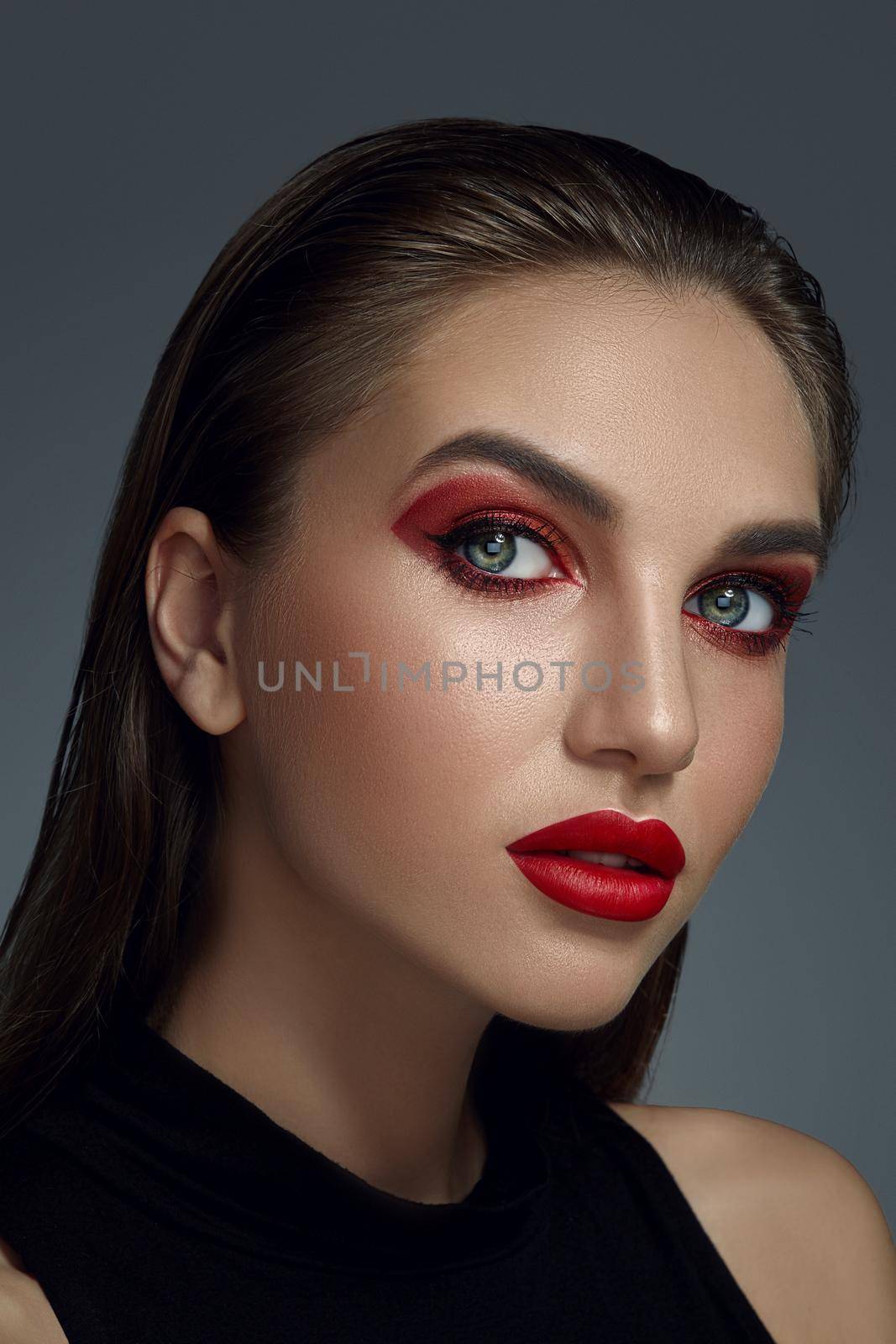 Close-up portrait of a female in black blouse looking at you while posing against a gray background. Professional make-up for gorgeous fashion model with an expressive eyes. Red lipstick and smokey eyes. Perfect skin.