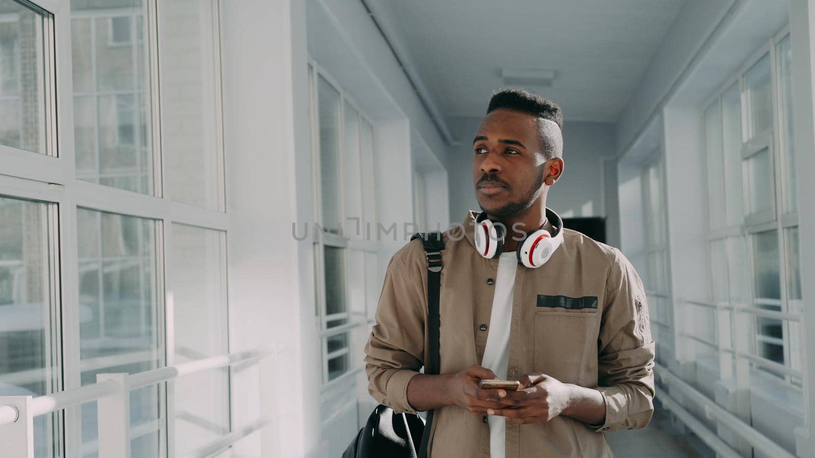 Handsome african american male student with big white headphones walking in long lighty corridor of college holding smartphone texting someone by silverkblack