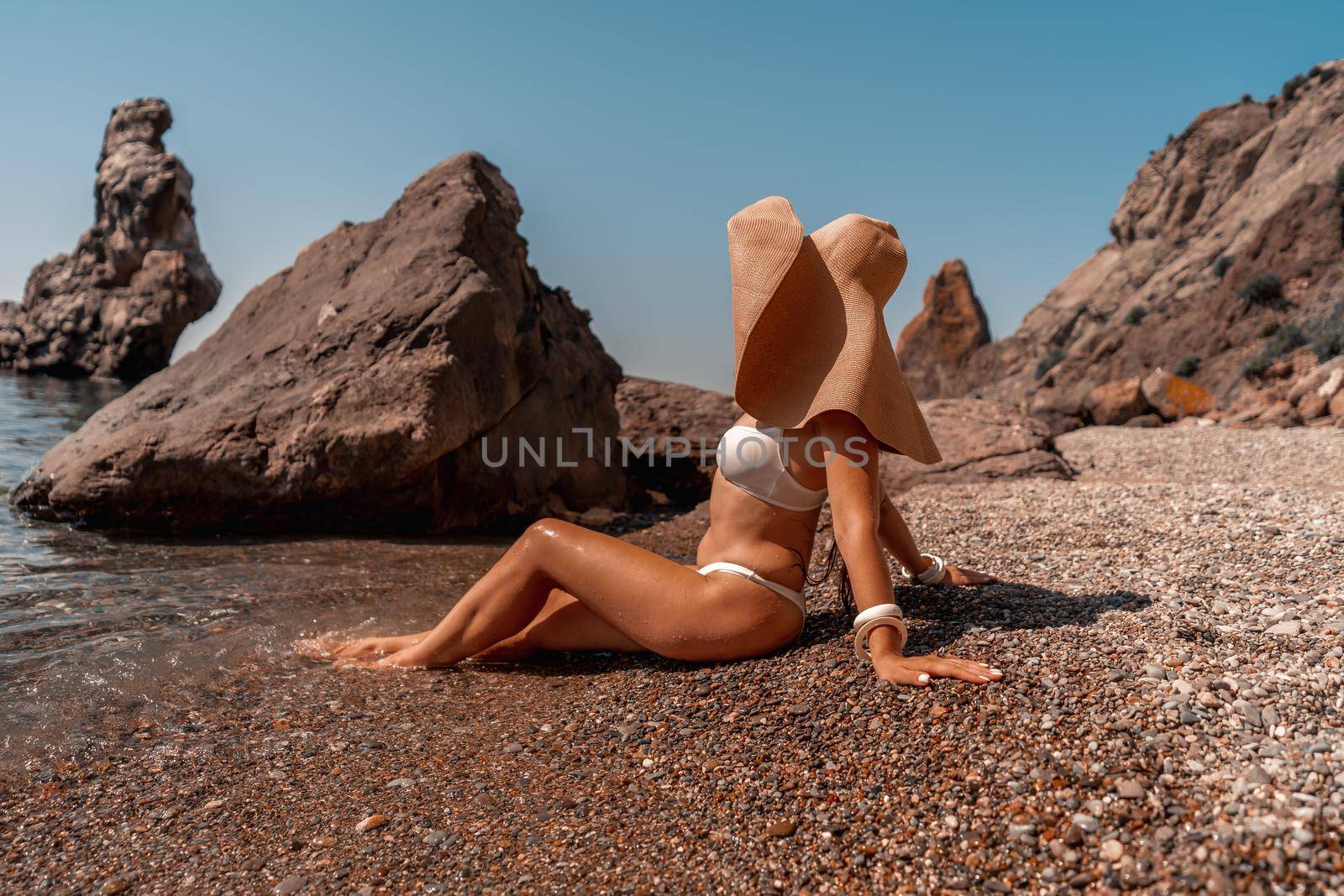 Tanned middle-aged woman with long hair and a white bathing suit. He sits on the seashore in a large sun hat with his back and looks at the sea