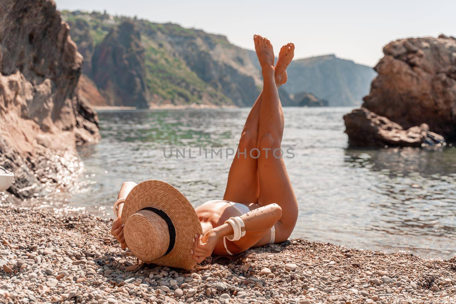 A young woman lies on the beach with her legs pointing to the sky, covering her body with a wide-brimmed straw hat. She is sunbathing on the ocean. The concept of vacation, travel, vacation