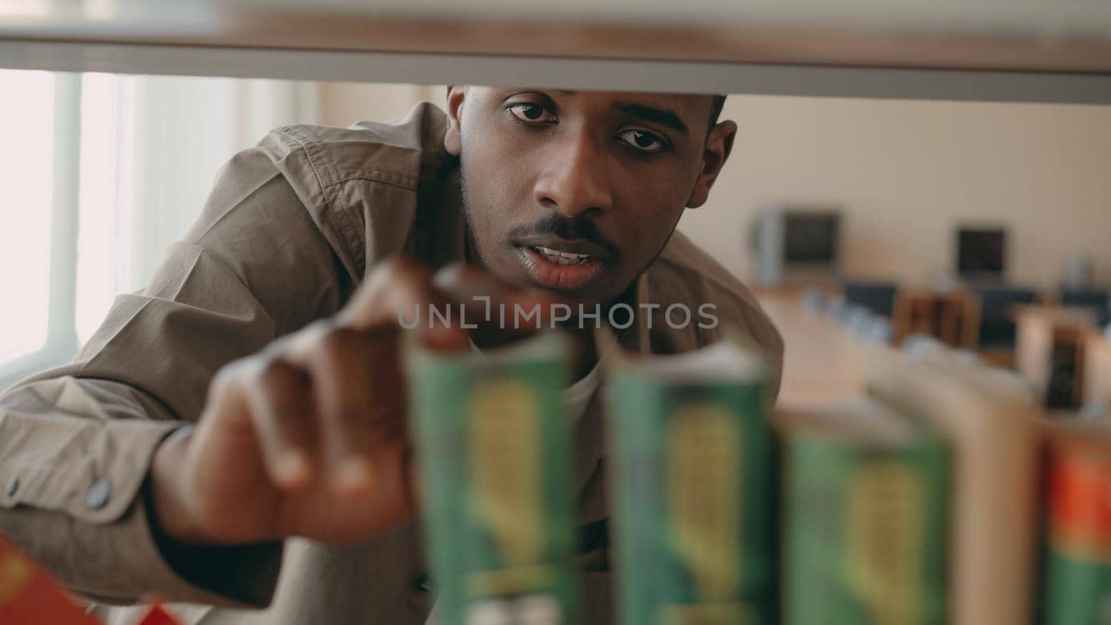 Afrcan-american serious handsome male student is looking for textbook on bookshelf in big library taking one book