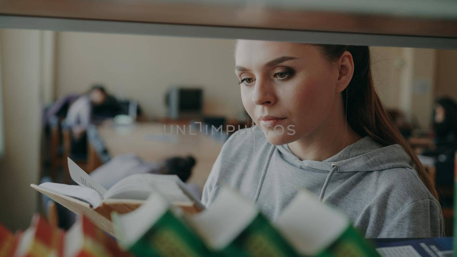 Young caucasian beautiful female student is standing near shelf with books in big lighty spacious library holding one, turning over pages and reading seriously