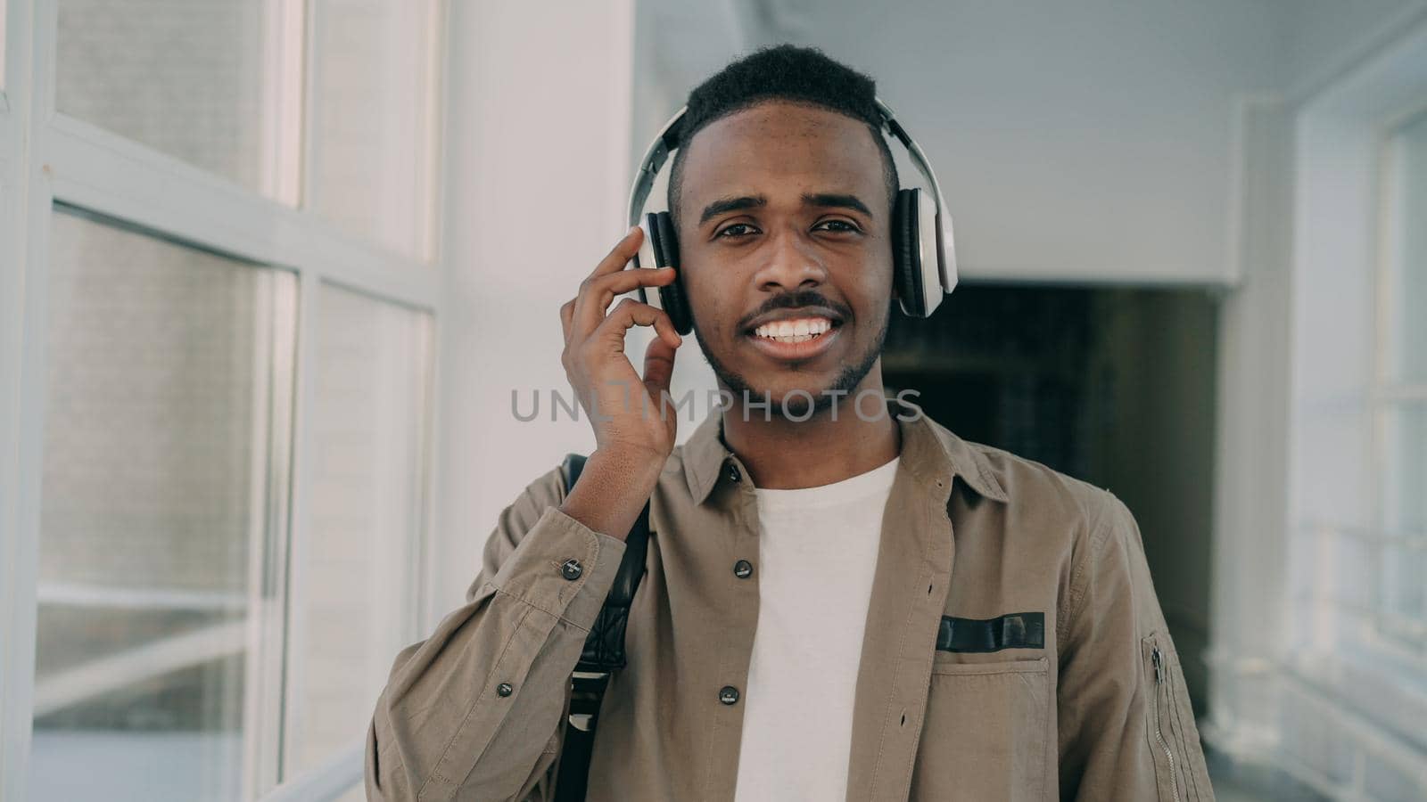 Portrait of young handsome student of african-american ethnicity standing in wide white spacious corridor of college with big headphones on his neck looking at camera and smiling.