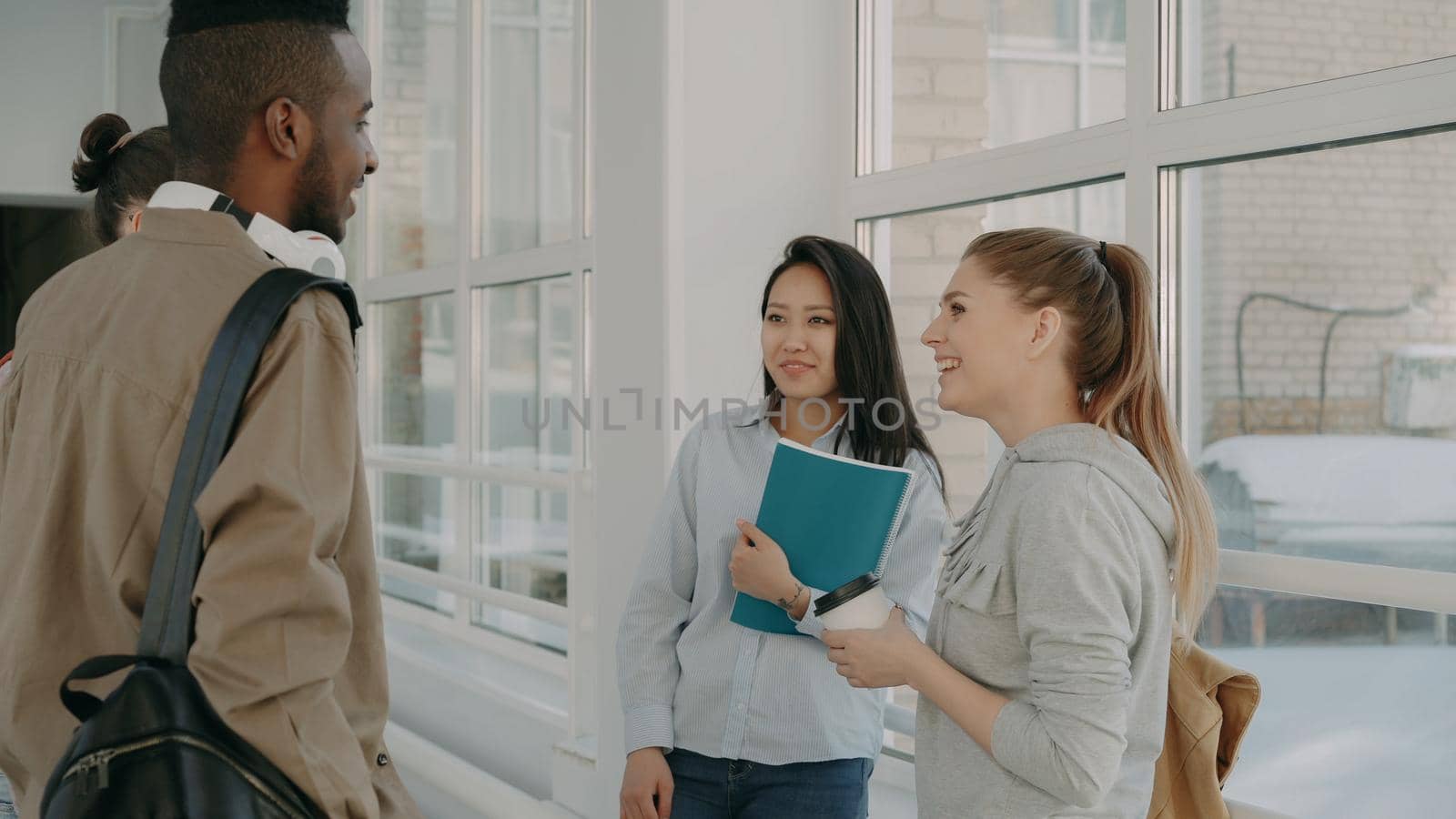 Multi-ethnic group of four students standing in wide lighty glassy hall in college talking to each other in positive way smiling ant laughing dinking coffee by silverkblack