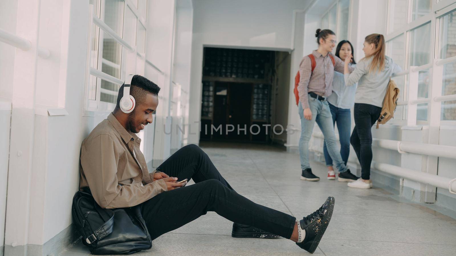 Young handsome african american student sitting on floor in white corridor with headphones on head listening to music holding smartphone texting someone. His classmates are behind him by silverkblack