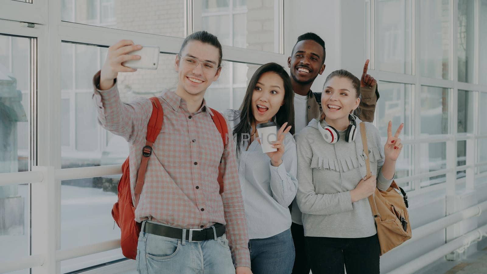Group of four multi-ethnic positive male and female students are standing in wide corridor. Hipster gut is holding smartphone making selfie of them all by silverkblack