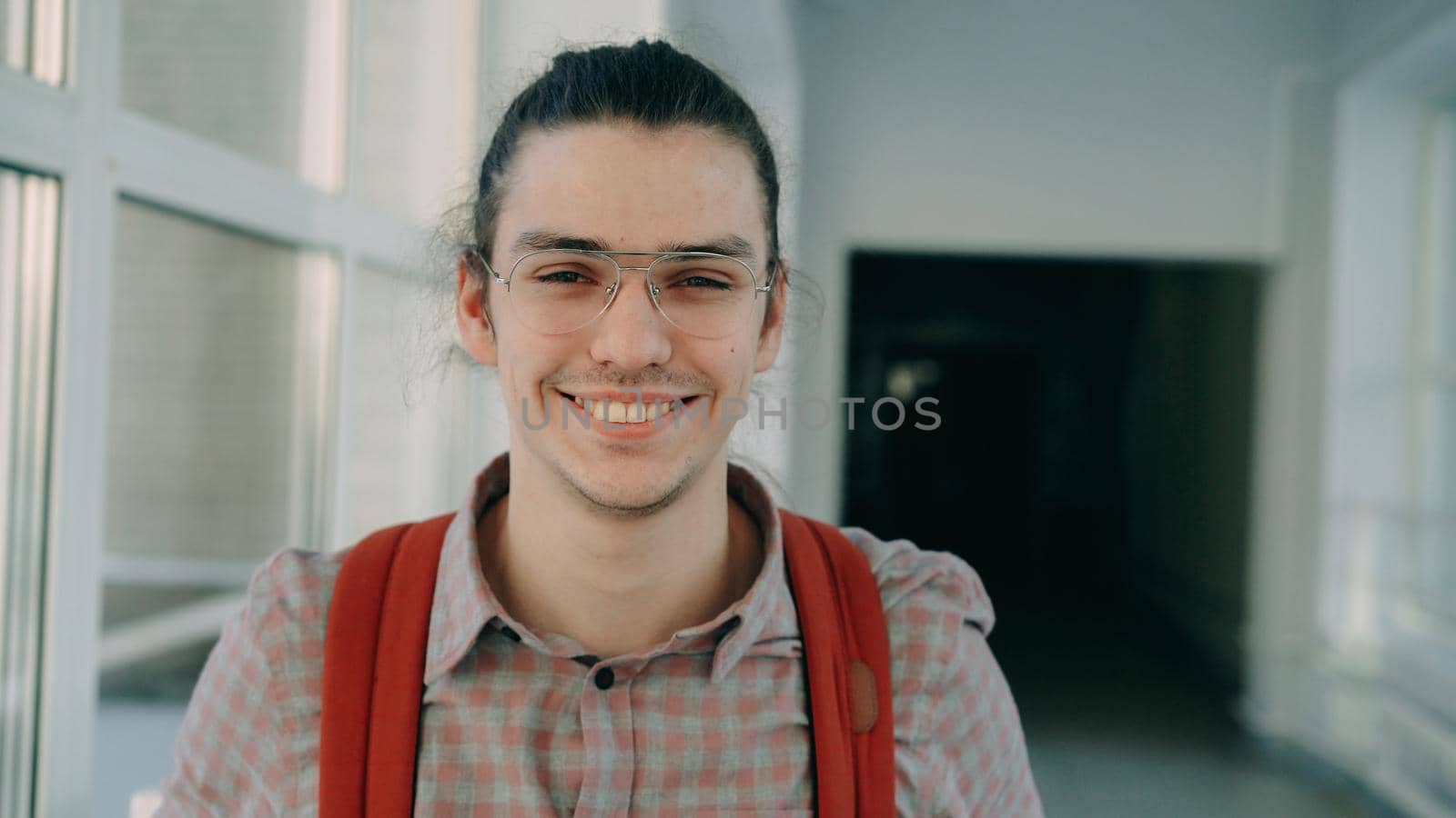 Portrait photo of young handsome attractive male student of caucasian ethnicity wearing glasses standing in wide white hallway indoors looking at camera and smiling positively