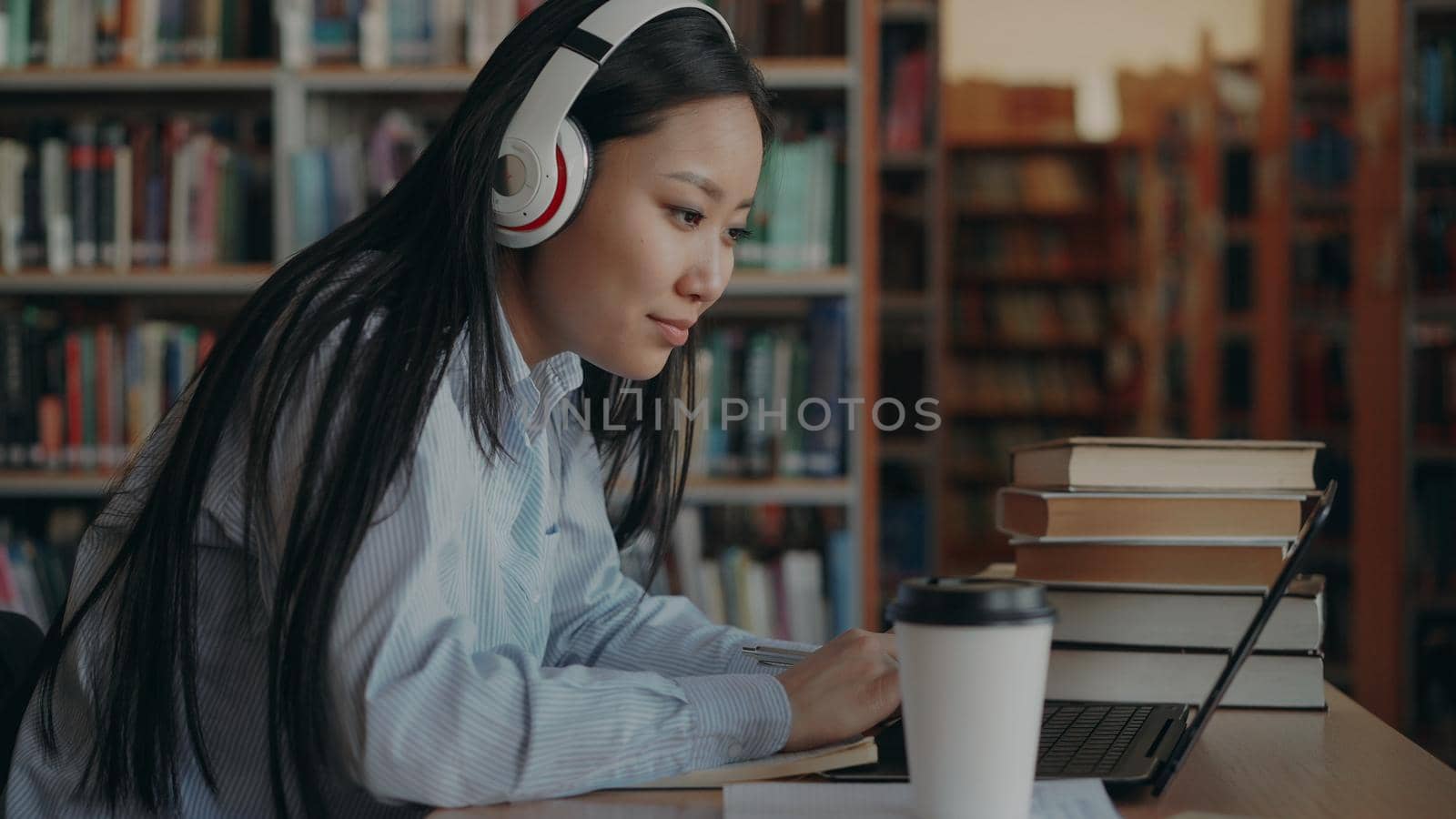 Attractive asian female student in headphones sitting at desk in library working concentrated at school project