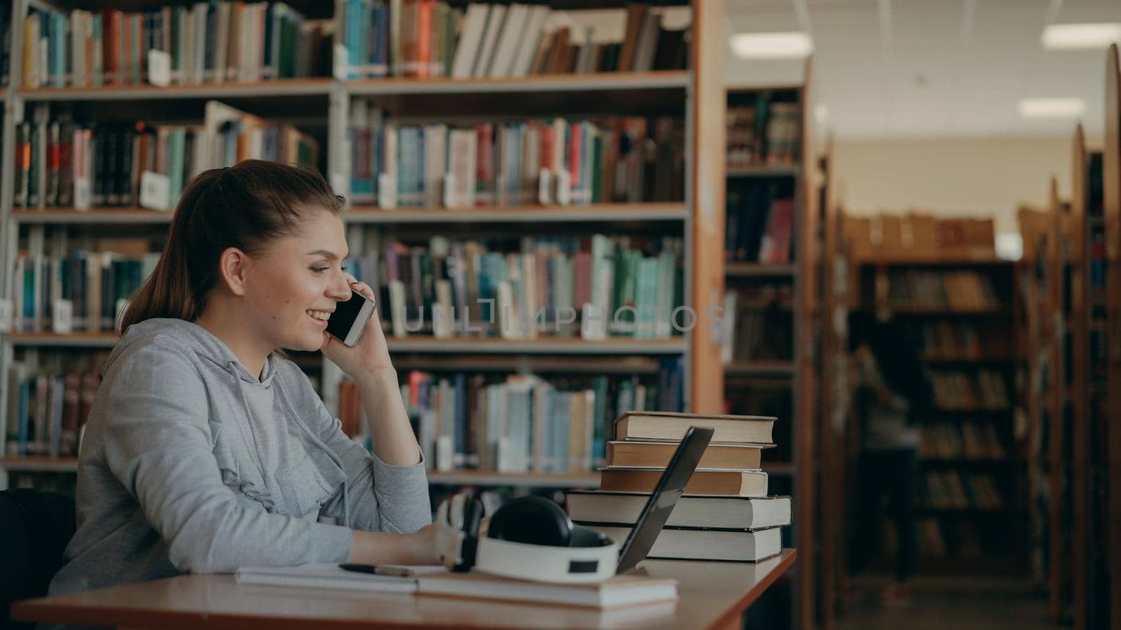 Attractive student girl in casual clothing is sitting at table using laptop computer in big university library talking on smartphone in positive way by silverkblack