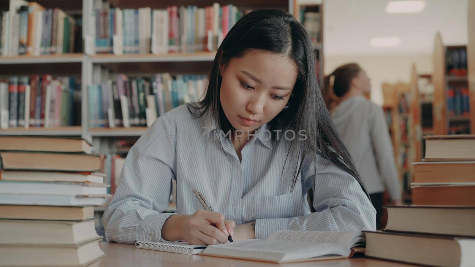 Young beautiful asian female student sitting at table surrounded by piles of books in lighty library rewriting text from textbook getting ready for important exam