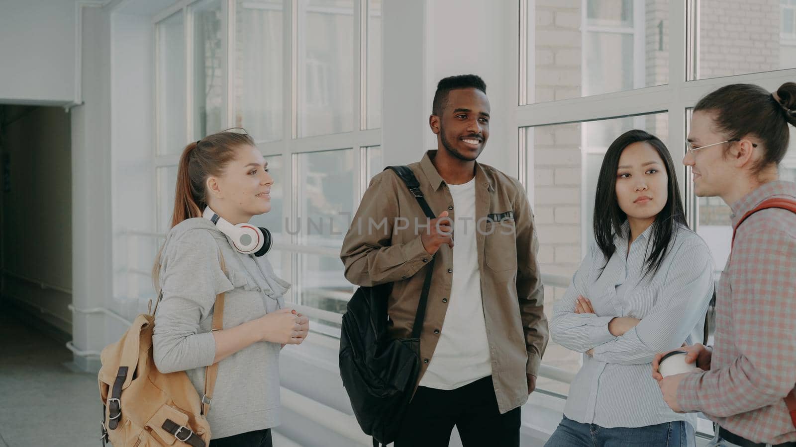 Young beautiful caucasian female student is standing in front of her three multi-ethnic groupmates discussing something together in positive way near window by silverkblack