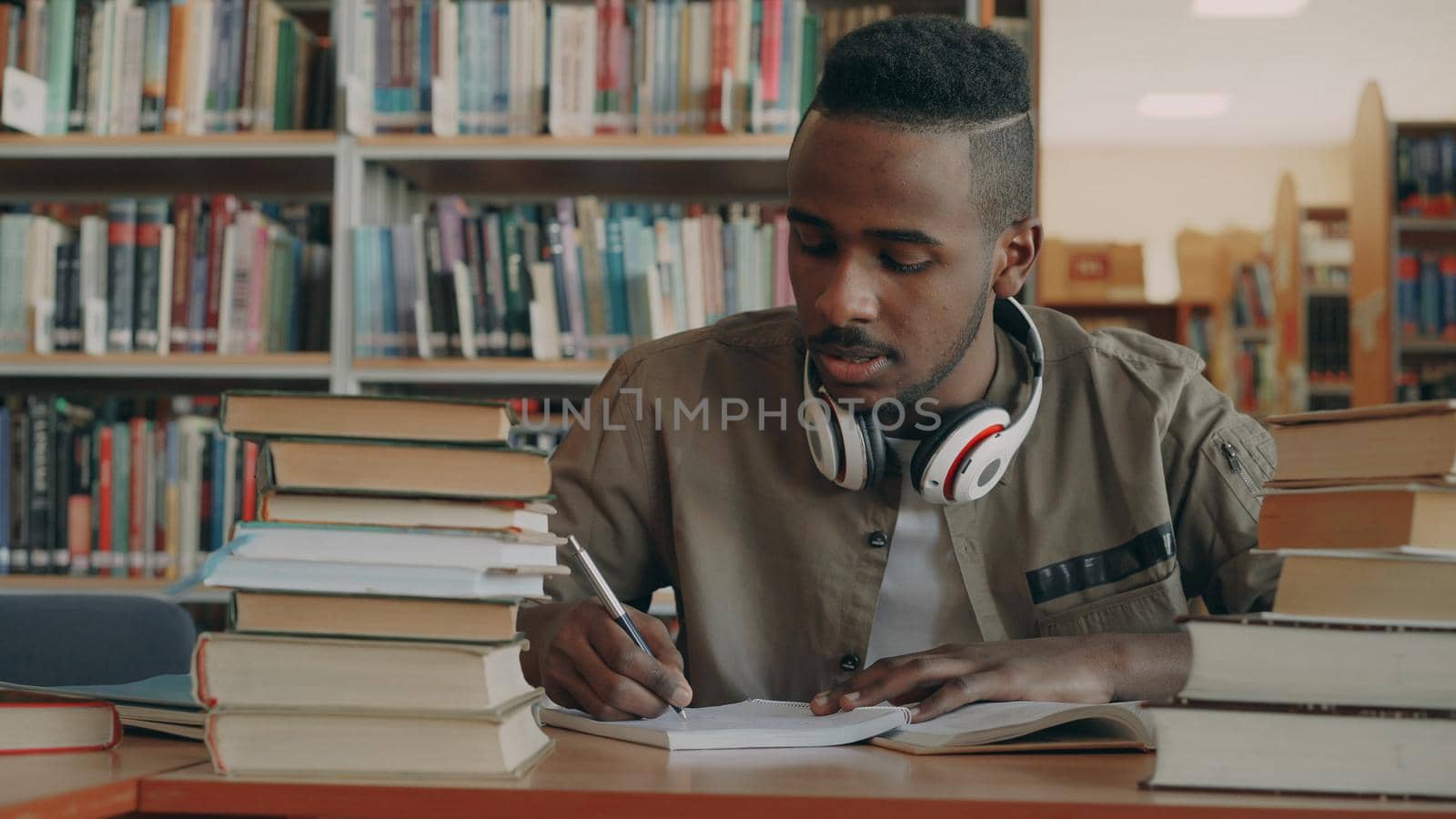 Positive african american young handsome guy with big headphones is sitting at table with books and writing in his copybook by silverkblack
