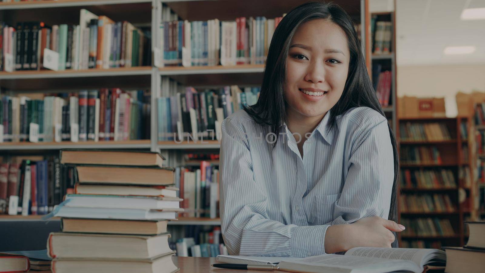 Portrait of young beautiful asian female student sitting at table with huge piles of textbooks and copybook in front of her in library looking at camera. She is smiling positively.