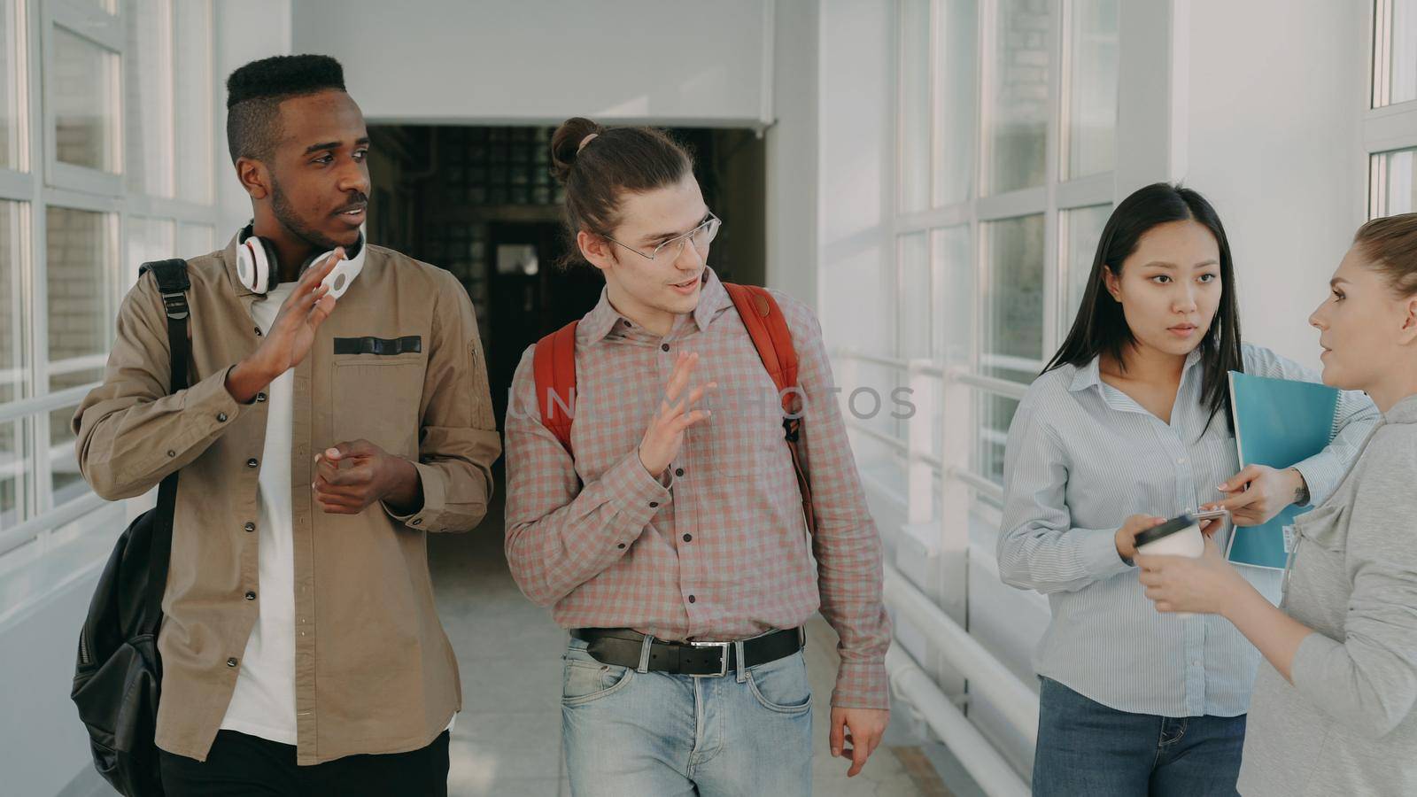 Two young handsome multi-ethnic friends walking down white glassy corridor in college talking passing by standing girls and waving hands to them by silverkblack