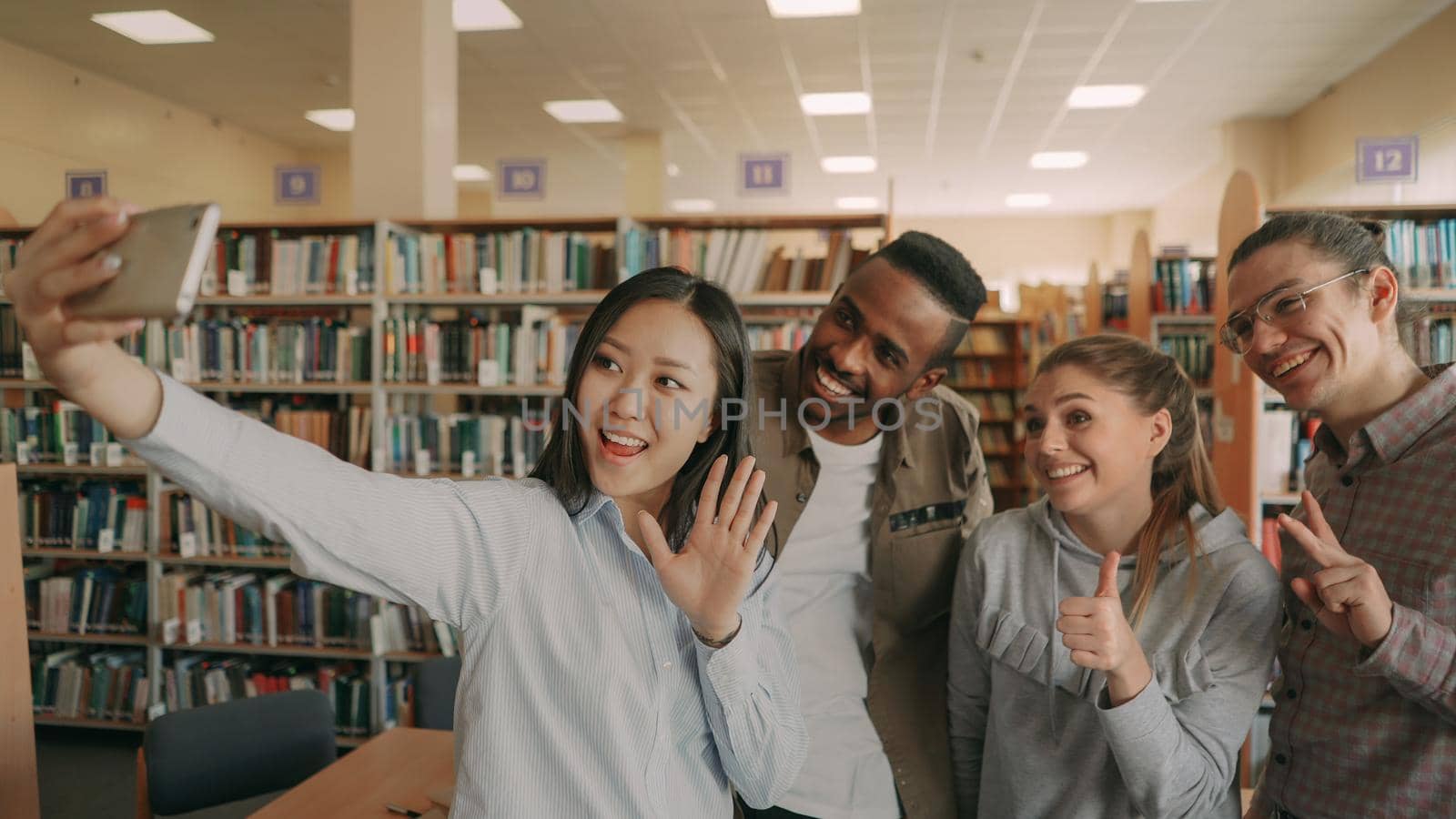 Group of international students have fun smiling and making selfie photos on smartphone camera at university library indoors. Cheerful friends have rest while preapre project together