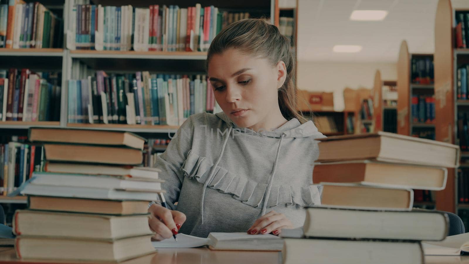 Concentrated young female student reading books and writing in notebook during working at school project in university library indoors