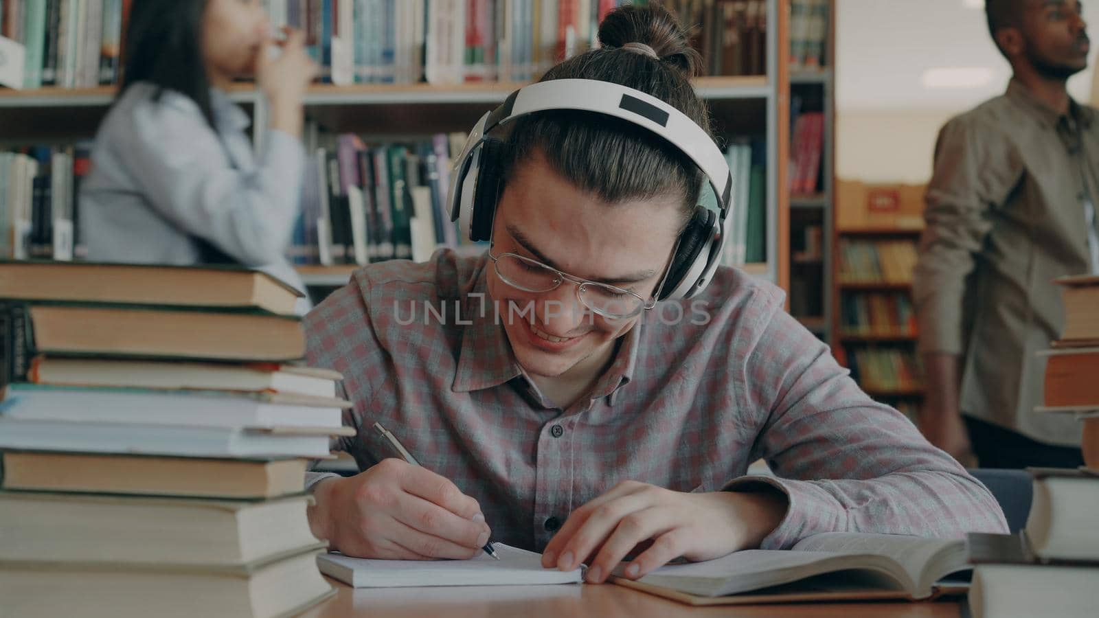 Young handsome caucasian schoolboy sitting at table in big college library is writing something in copybook. He is smiling and looks positive and happy, books lying on table
