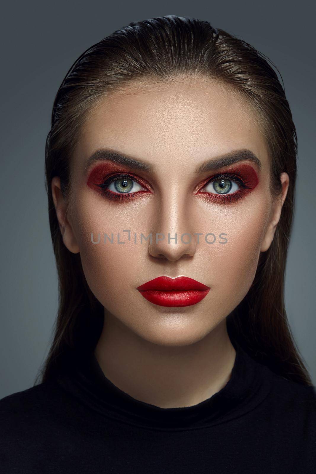 Close-up portrait of a girl in black blouse looking at the camera and posing against a gray background. Professional make-up for gorgeous fashion model with an expressive eyes. Red lipstick and smokey eyes. Perfect skin.