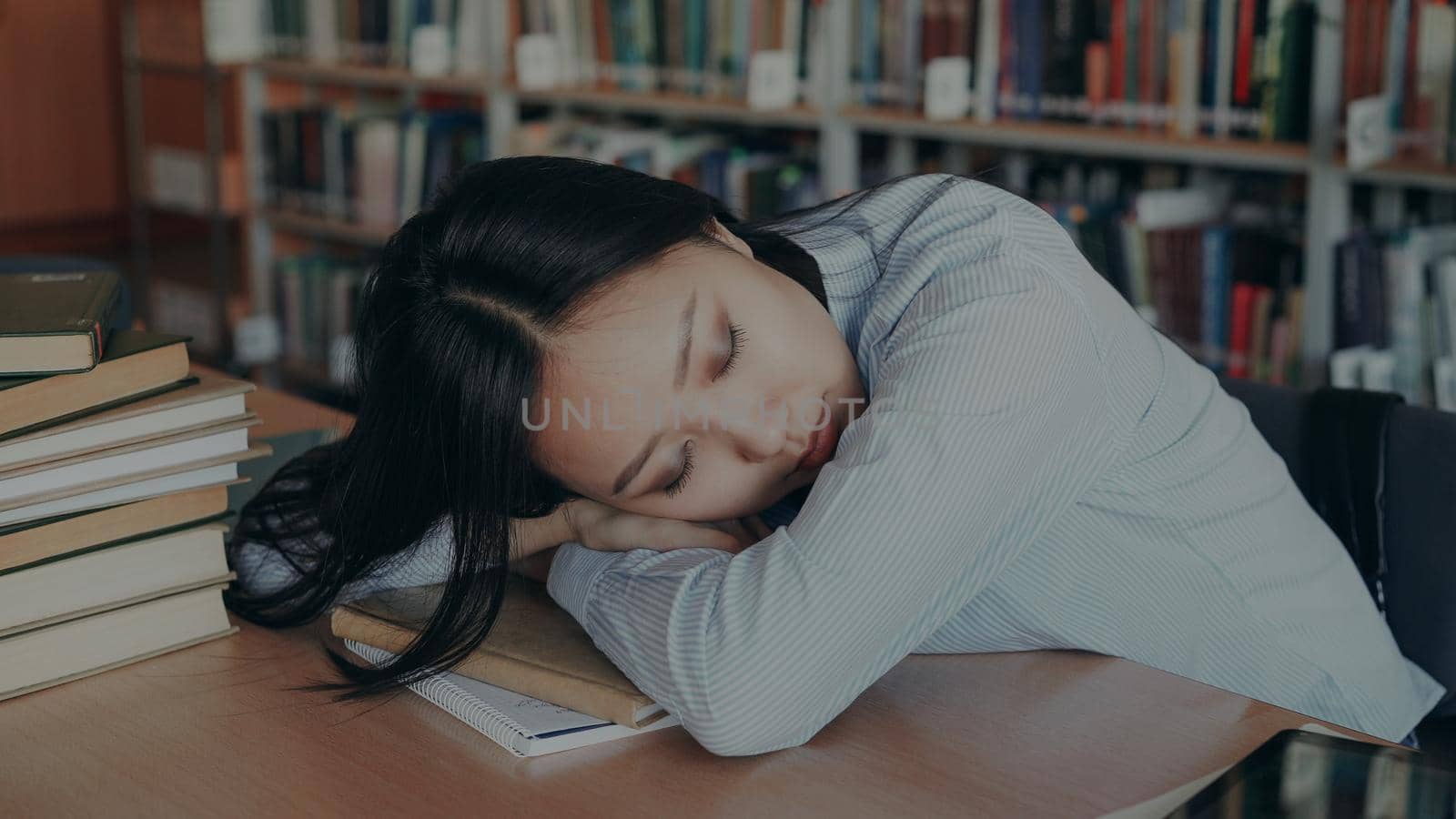Young beautiful tired asian female student is sitting at desk with her head on it, she is sleeping on textbook surrounded by piles in big university library by silverkblack