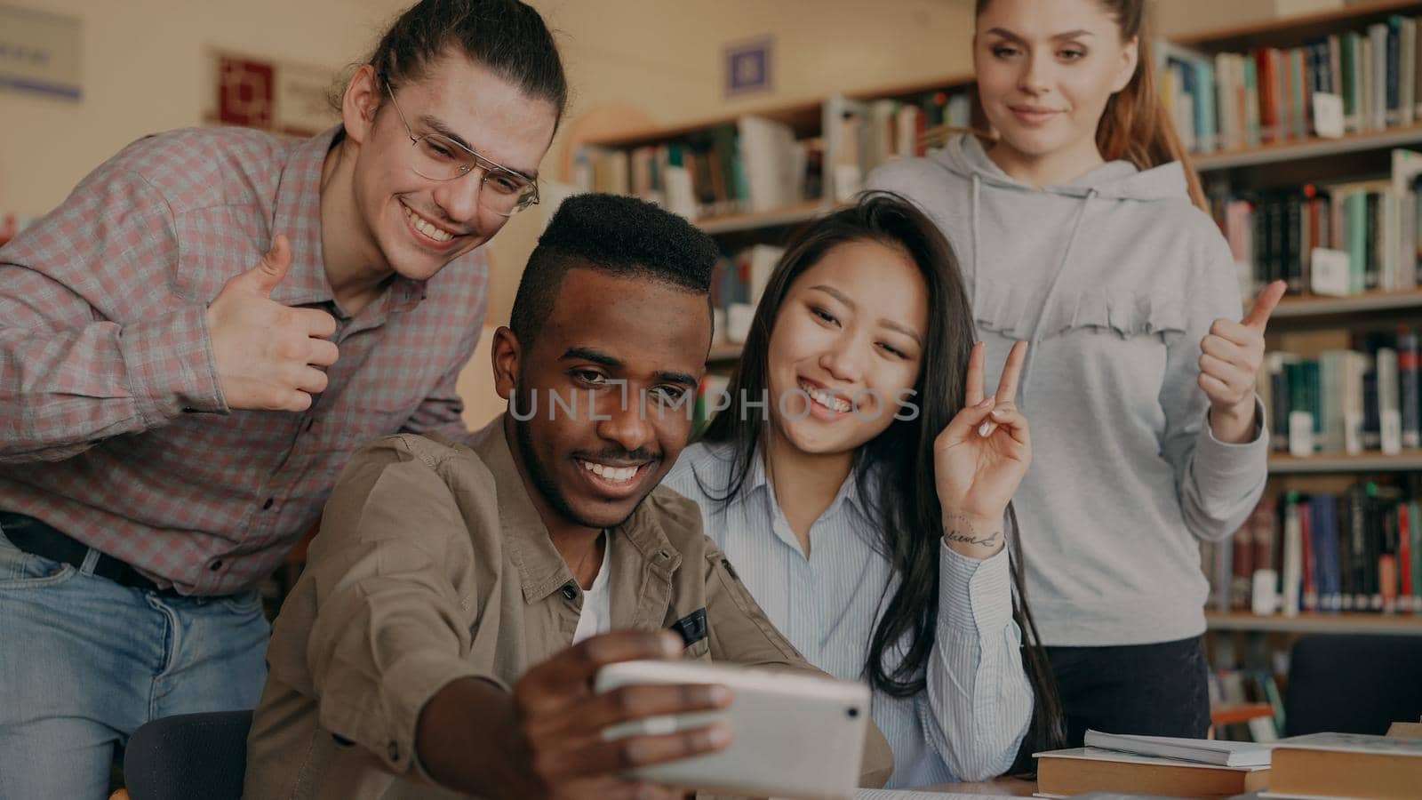 Group of international students have fun smiling and making selfie photos on smartphone camera at university library indoors. Cheerful friends have rest while preapre project together