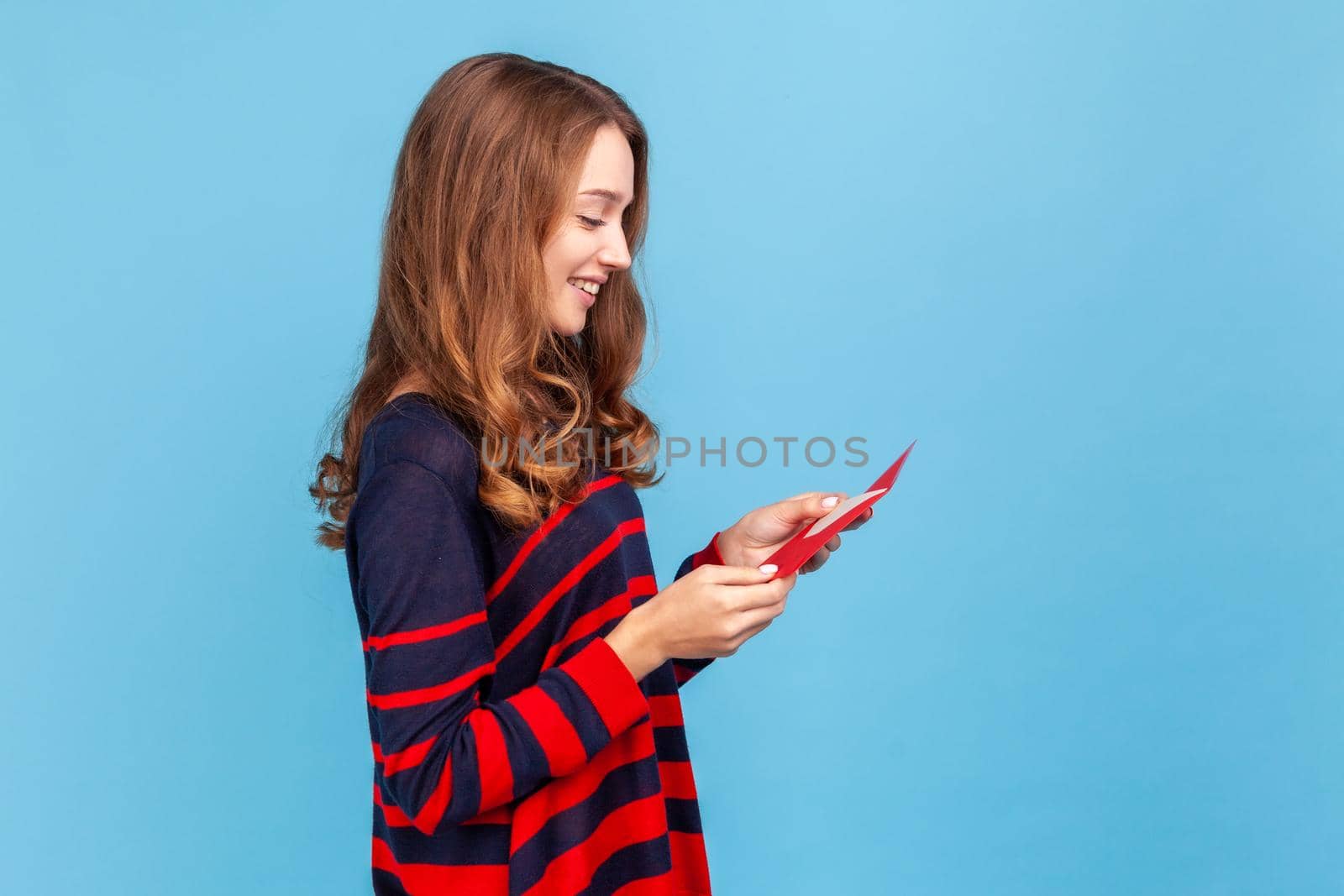 Side view portrait of woman in striped casual style sweater, reading letter from red envelope, gets greeting card, having positive romantic expression. Indoor studio shot isolated on blue background.