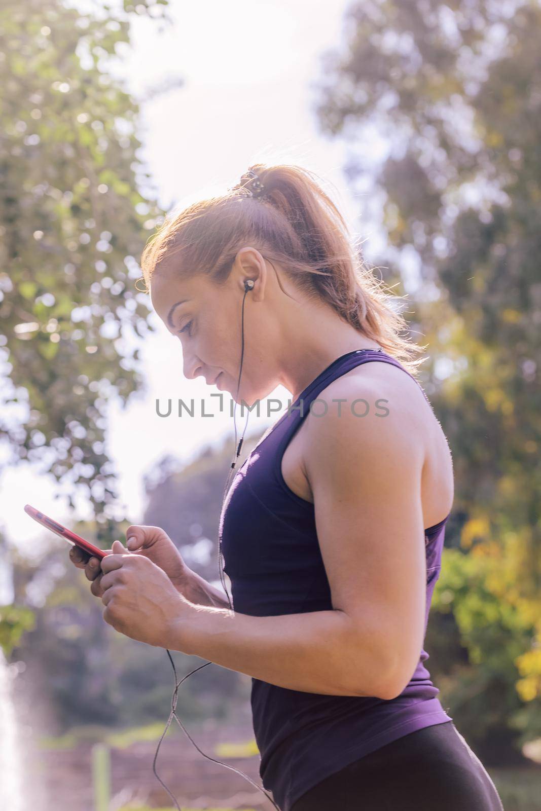 vertical photo of an attractive blonde sports woman checking her phone app before workout outdoors at park, fitness accessories
