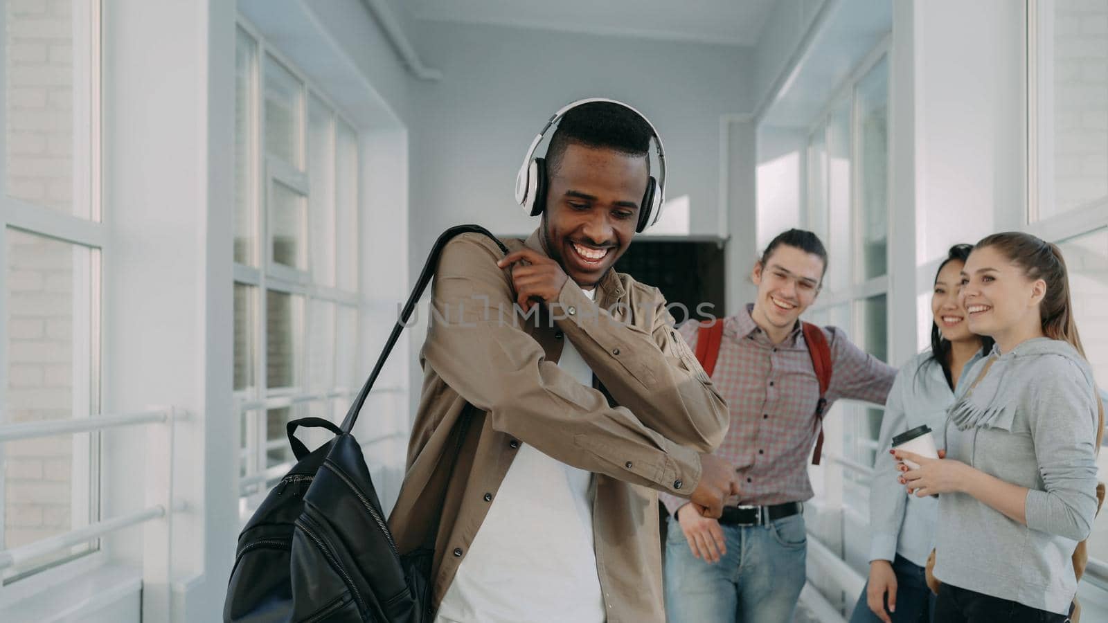 Young handsome african-american student is walking dancing in white corridor of college listening to music in big headphones by silverkblack