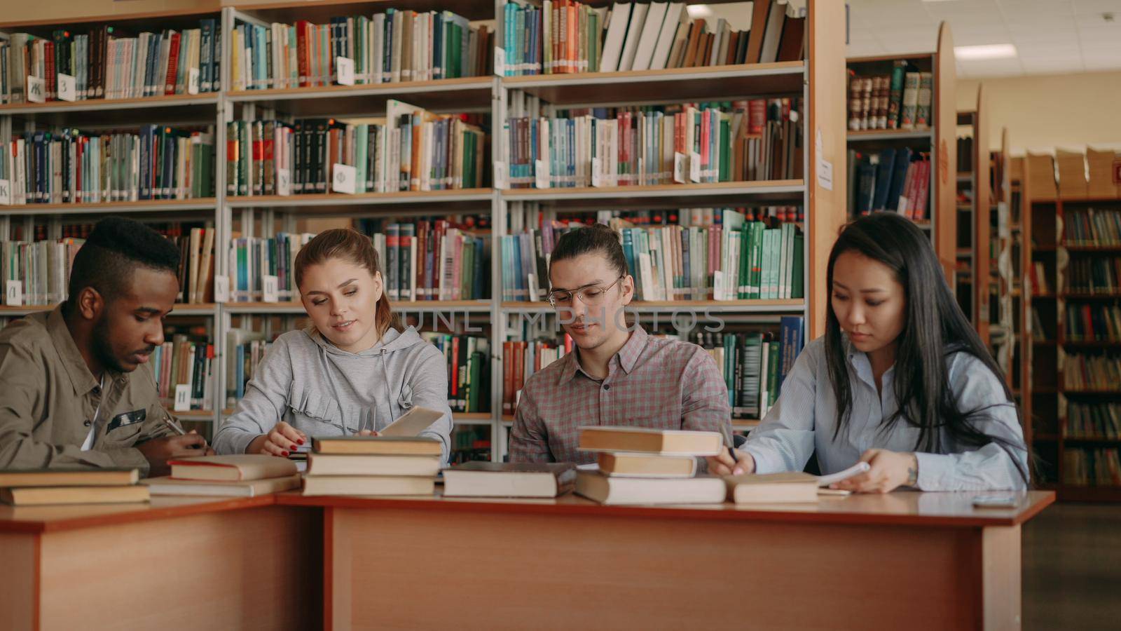 Multi ethnic group of students chatting and preparing for examination while sitting at the table in university library indoors