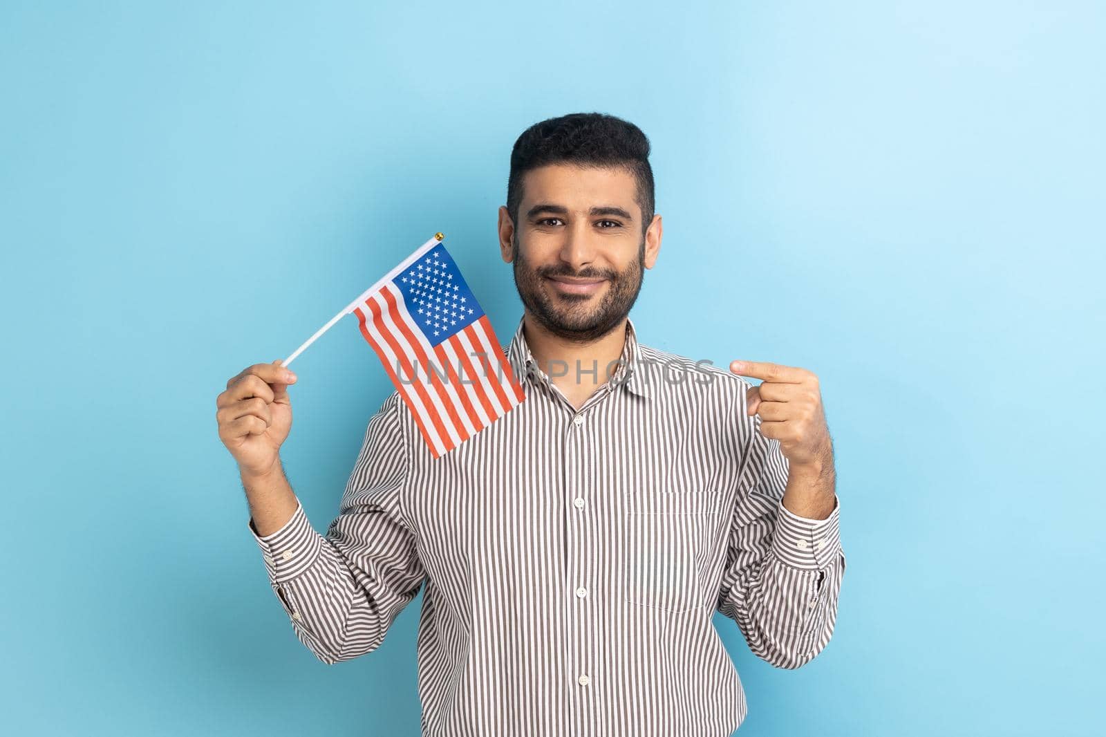 Portrait of happy positive man with beard standing and pointing at american flag, celebrating national holiday, wearing striped shirt. Indoor studio shot isolated on blue background.