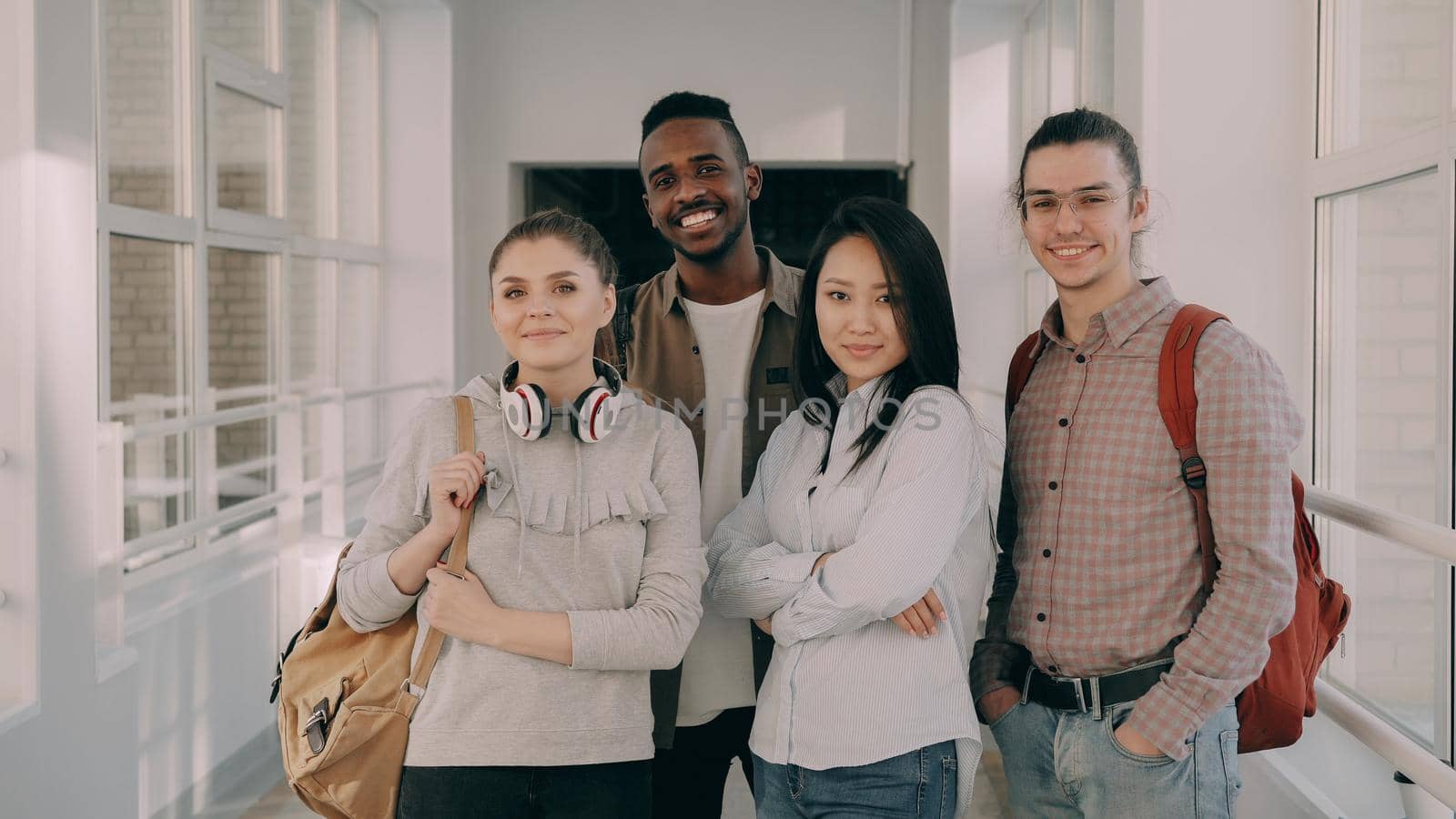 Portrait of four happy smiling positive attractive multi-ethnic male and female students standing motionlessly in spacious white wide corridor in university looking at camera