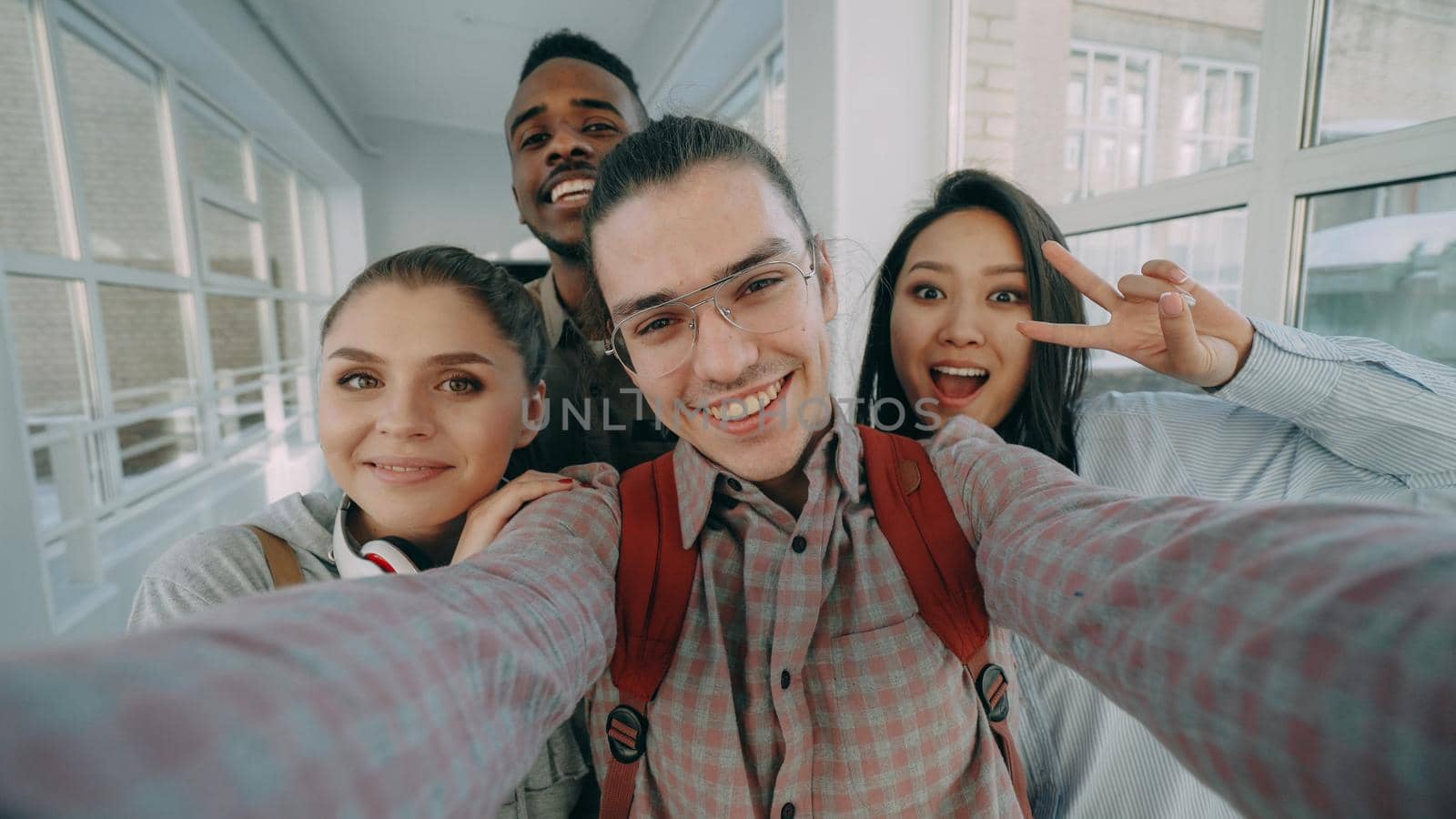 Point of view of four positive cheerful attractive multi-ethnic friends talking selfie photos holding smartphone and having fun laughing while standing in corridor of university.
