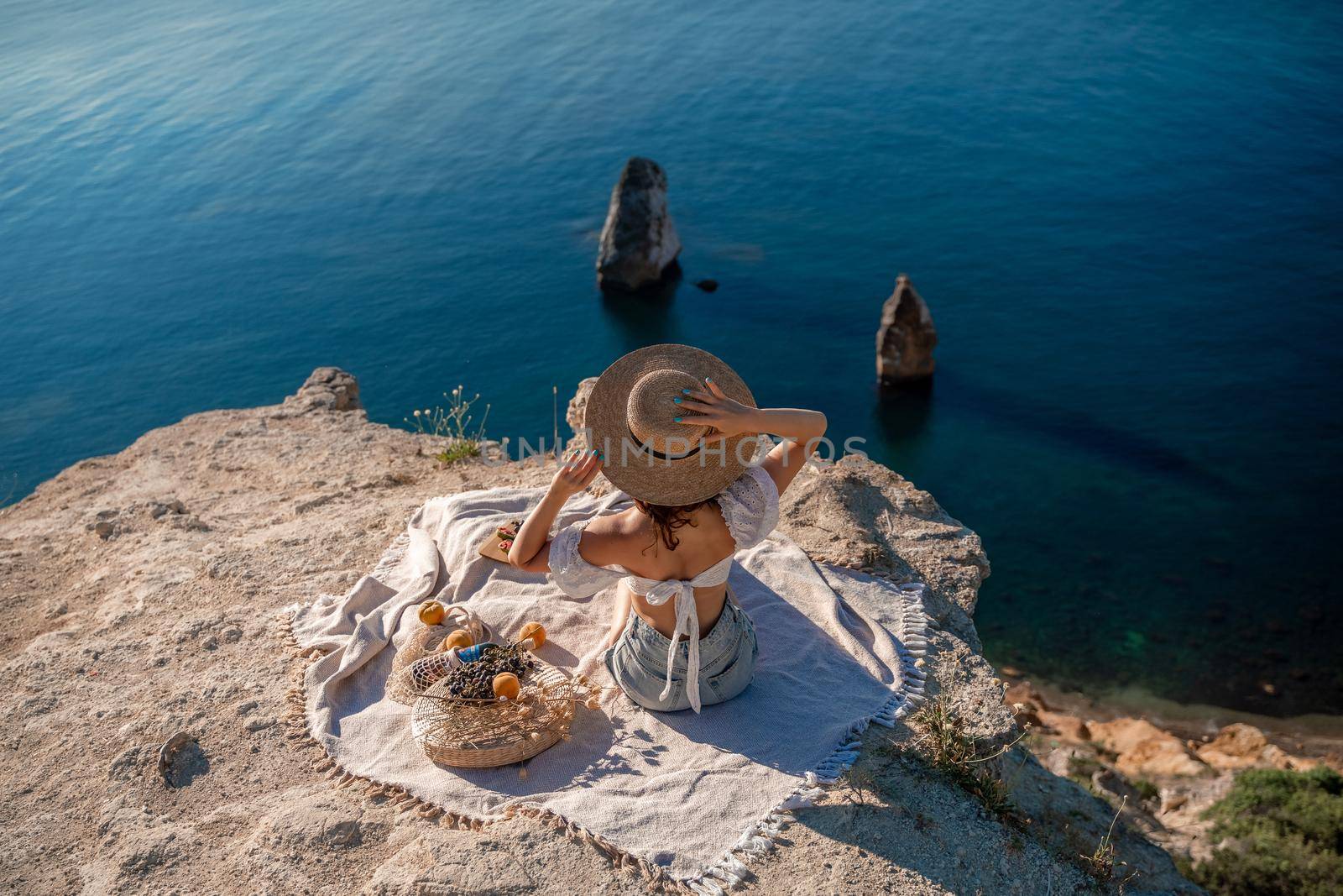 Street photo of a beautiful woman with dark hair in a white top, shorts and a hat having a picnic on a hill overlooking the sea.