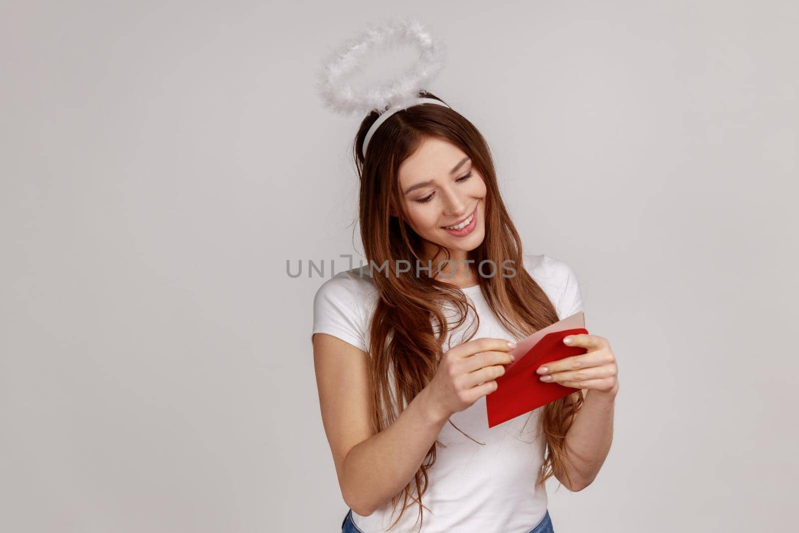 Portrait of charming pleased woman with nimbus over head holding red envelope, reading romantic letter with smile, wearing white T-shirt. Indoor studio shot isolated on gray background.