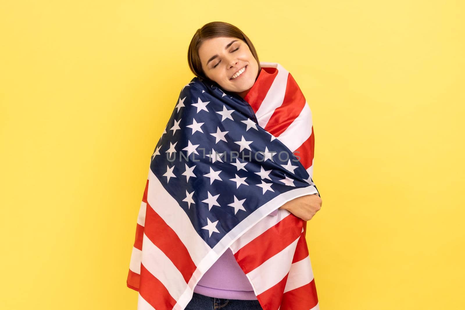 Portrait of kind friendly attractive woman standing wrapped in american flag, looking at camera with pleasant smile, wearing purple hoodie. Indoor studio shot isolated on yellow background.