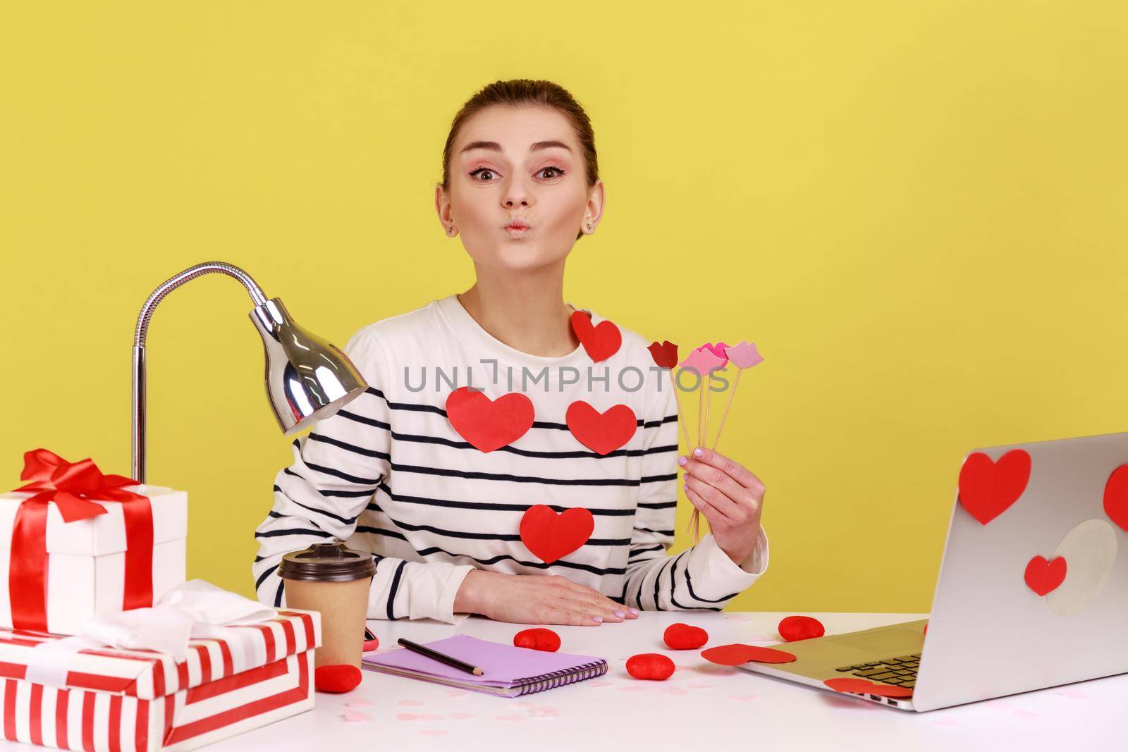Portrait of funny optimistic festive woman office manager sitting on workplace with pout lips and holding party props in hands. Indoor studio studio shot isolated on yellow background.
