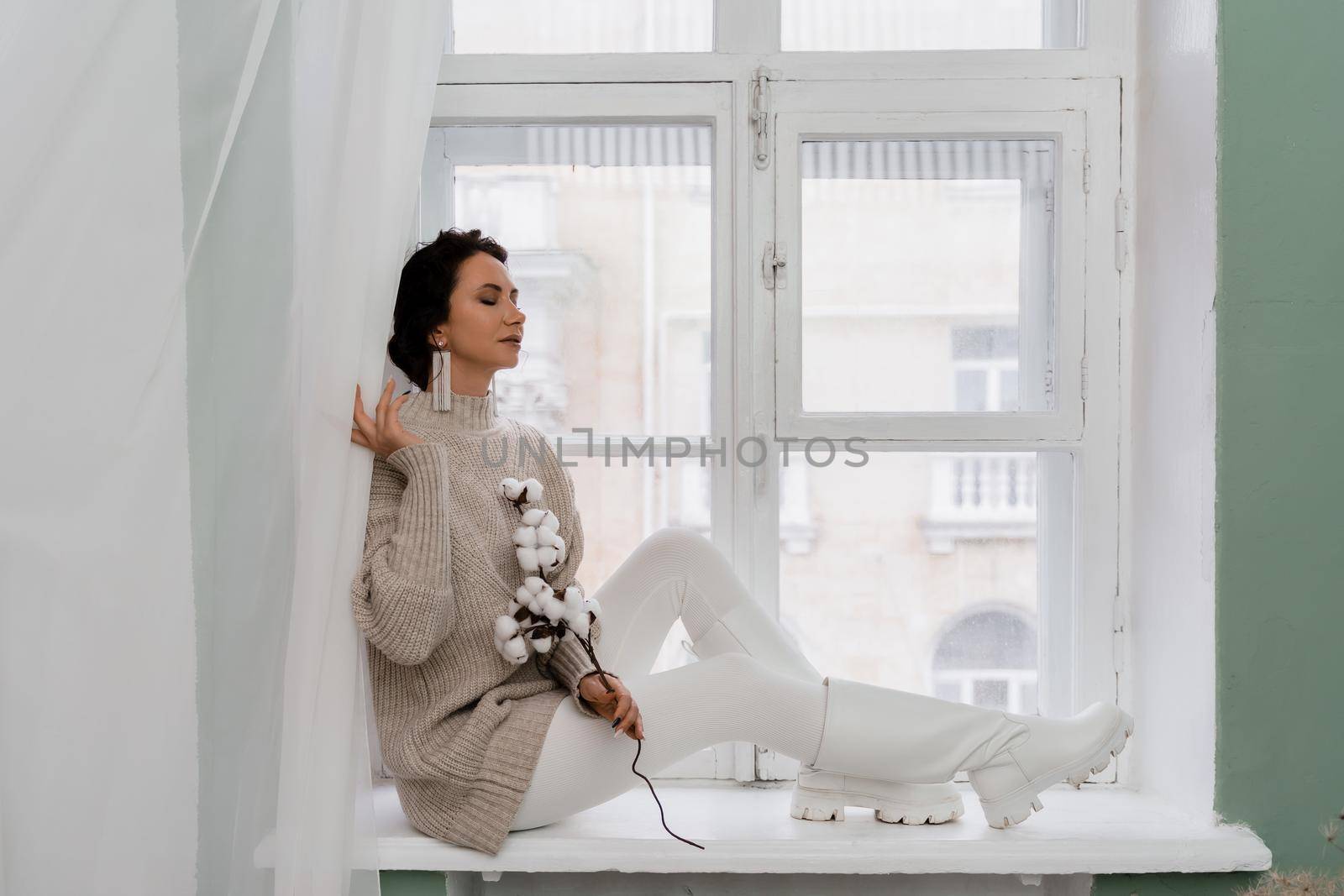 Free time for rest. Profile of a beautiful woman sitting on a white window sill at home, holding a cotton plant in her hands. She is wearing a sweater, leggings and white boots