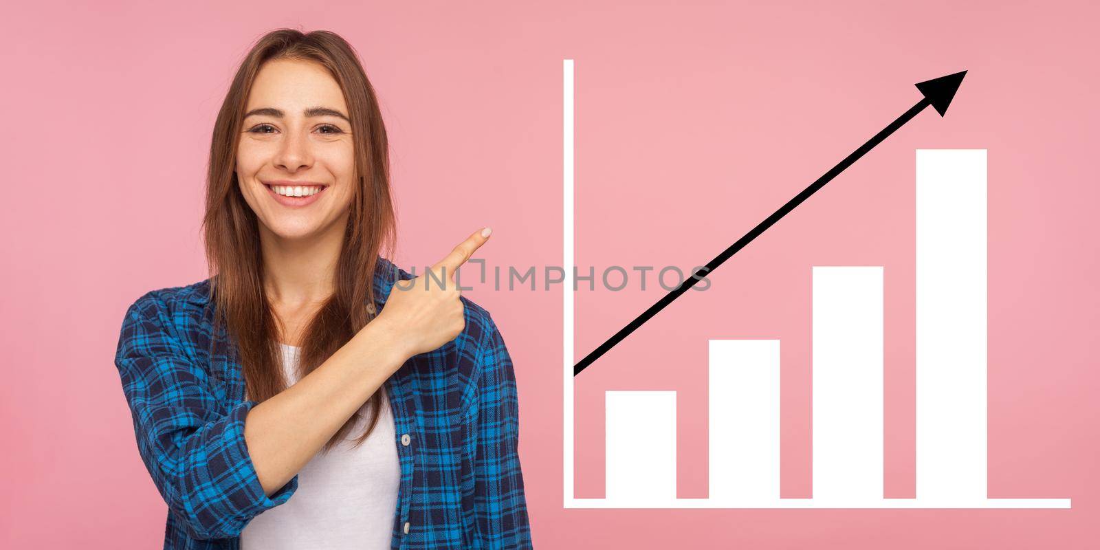 Portrait of happy joyful young woman standing, pointing aside and showing business growth graph. indoor studio shot isolated on pink background
